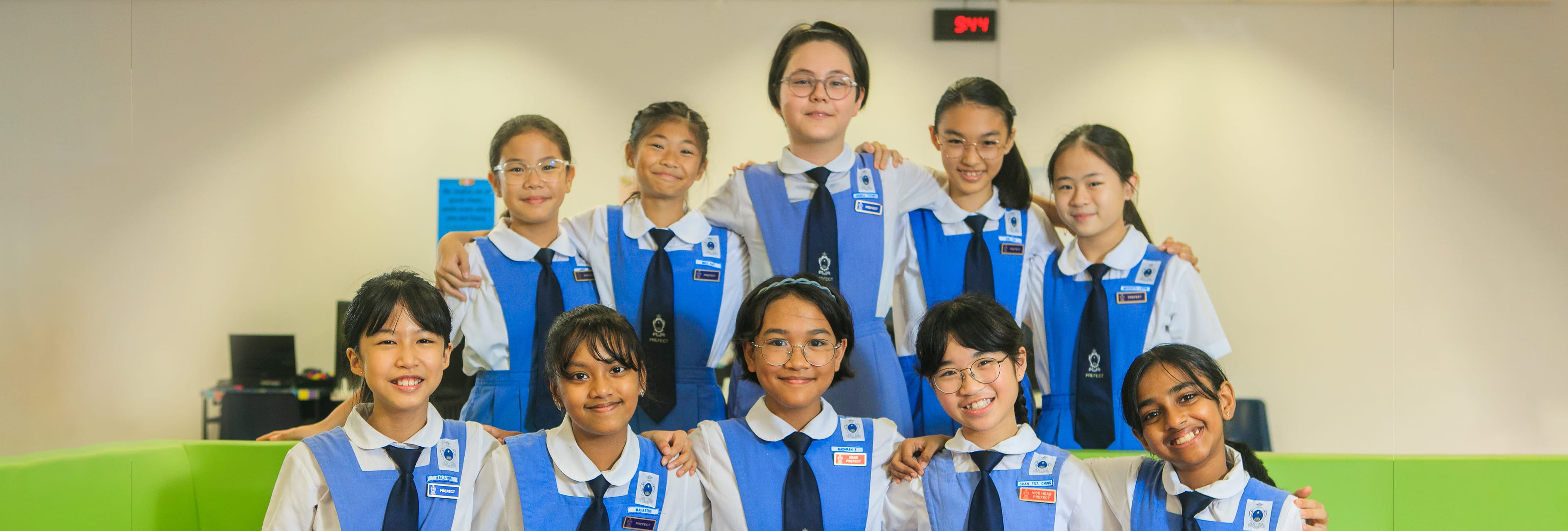 Ten smiling schoolgirls in blue and white uniforms with ties pose together in two rows indoors.