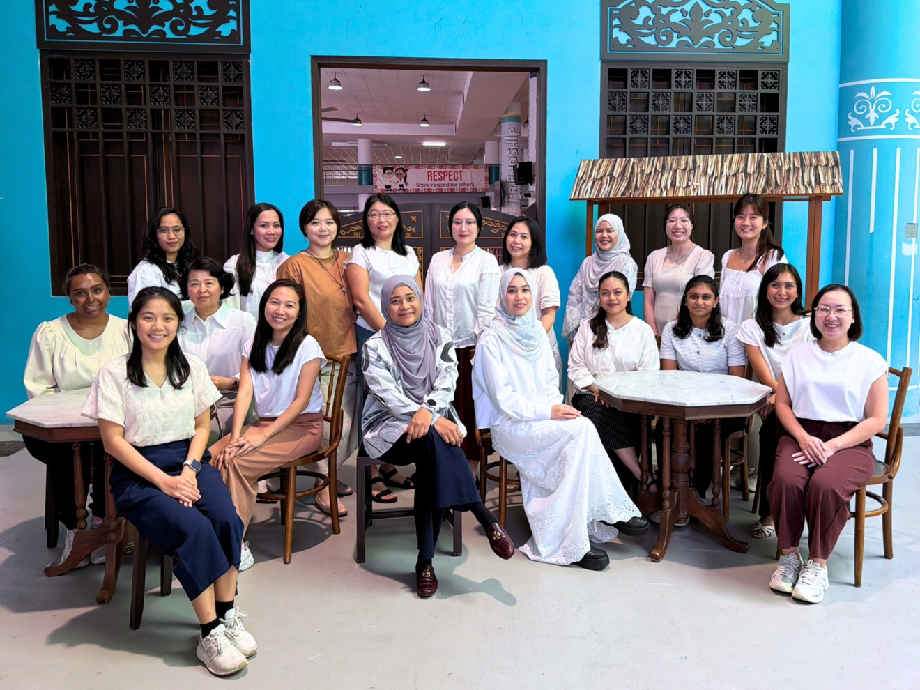 A group of about 18 women posing together in front of a bright turquoise building with wooden furniture.