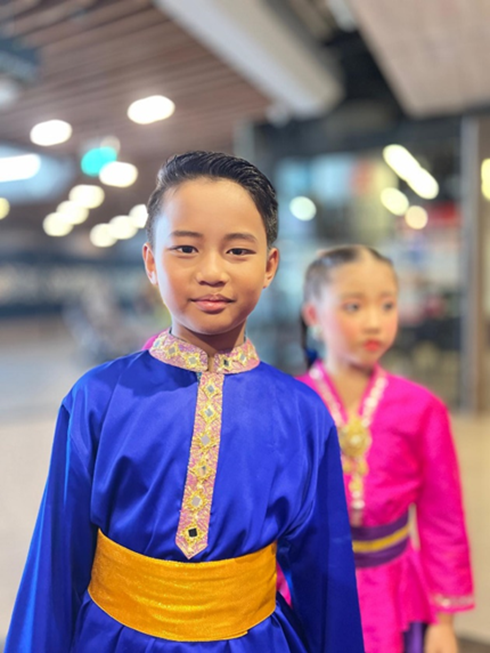 Two children in traditional Southeast Asian costumes, one in blue with gold sash, another in pink, indoors.