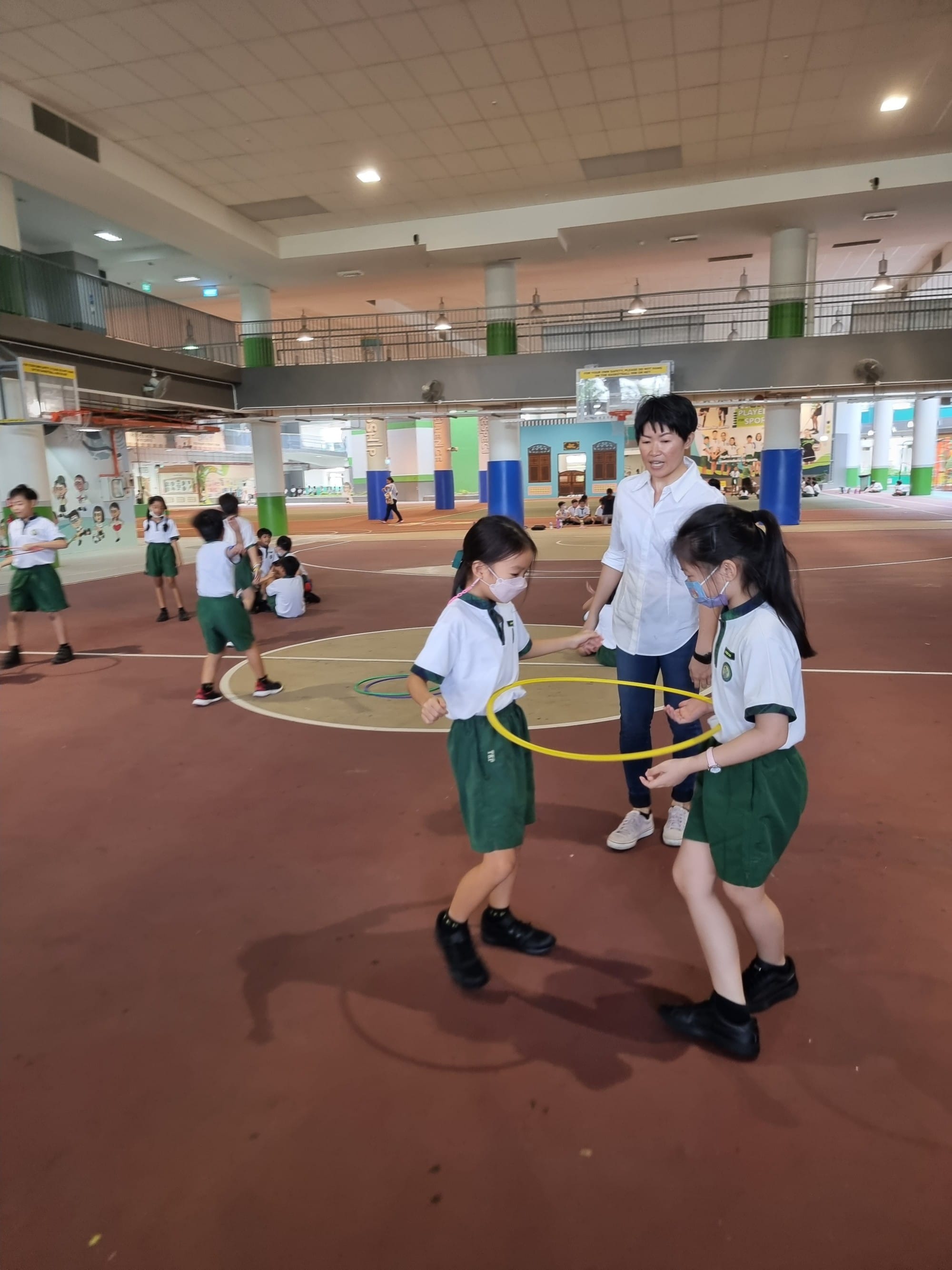 Two young students in green uniforms pass a yellow hula hoop while a teacher supervises in an indoor school hall.