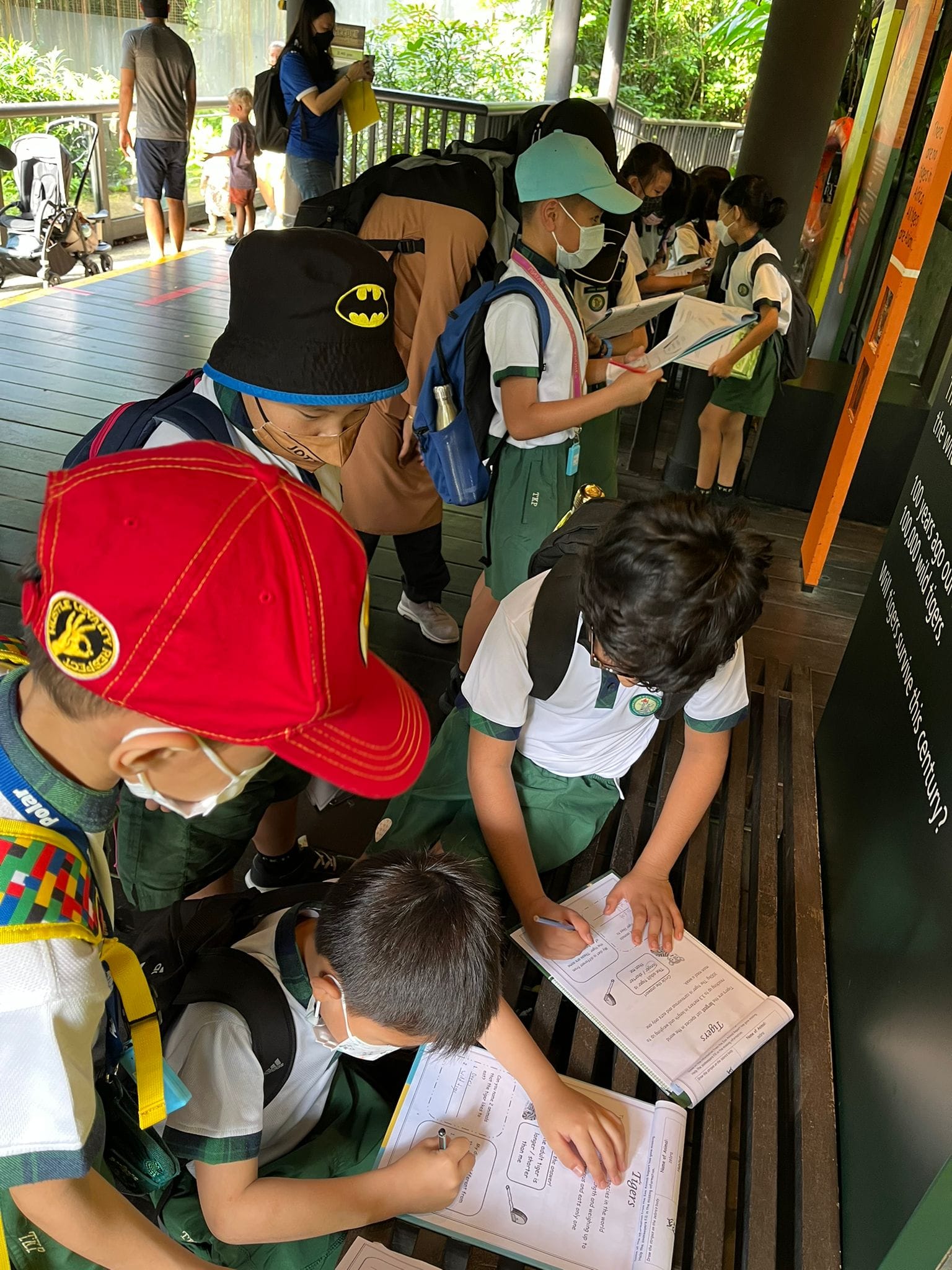 Children in school uniforms and masks fill out worksheets on a wooden walkway surrounded by greenery.