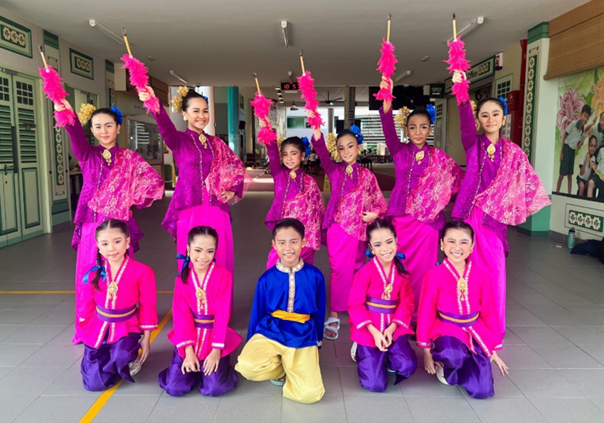 Group of children in bright pink and purple traditional costumes posing, some holding pink feathered props raised overhead.