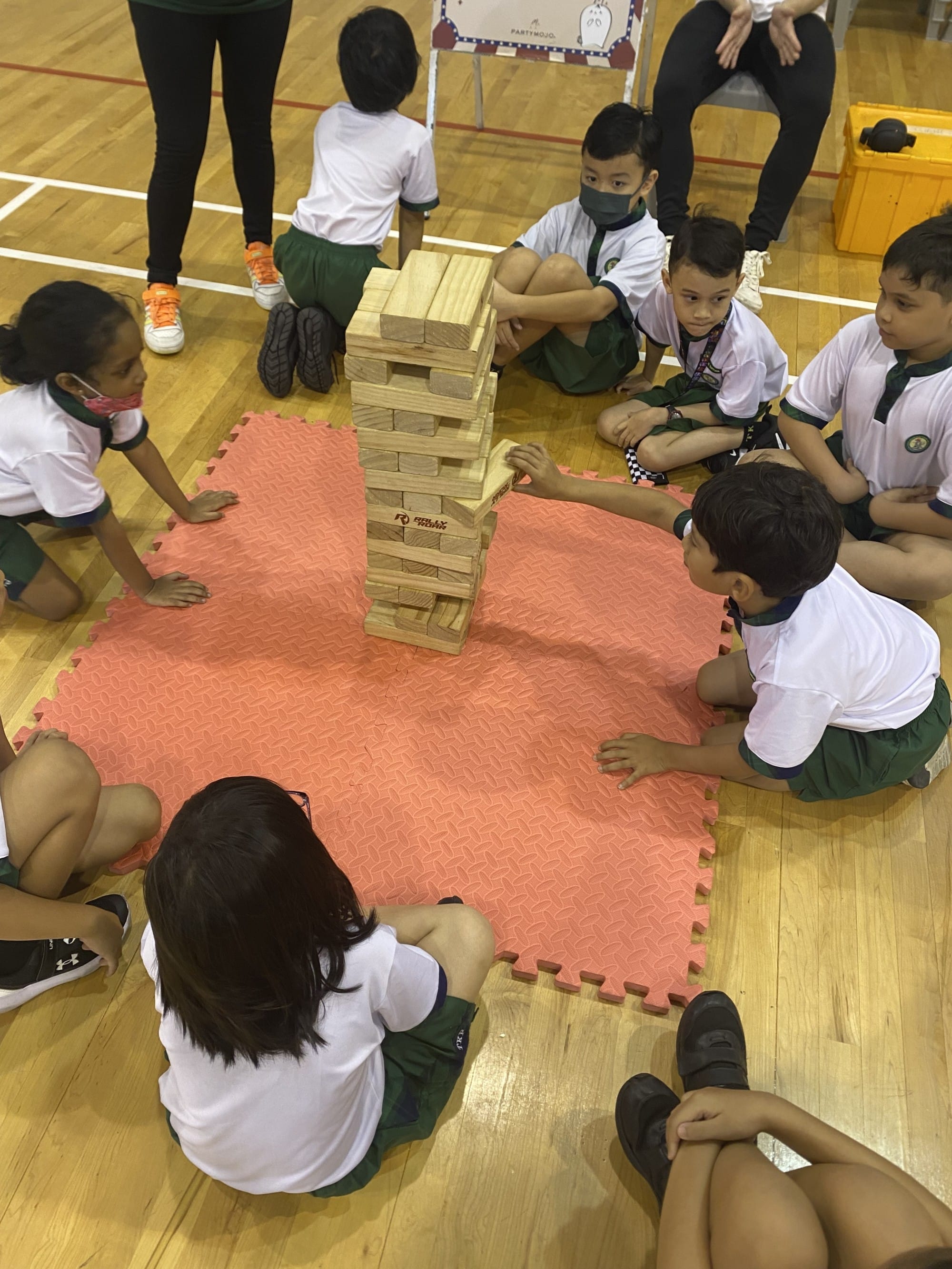 Children in school uniforms sit in a circle on a red foam mat playing a giant Jenga tower game indoors.