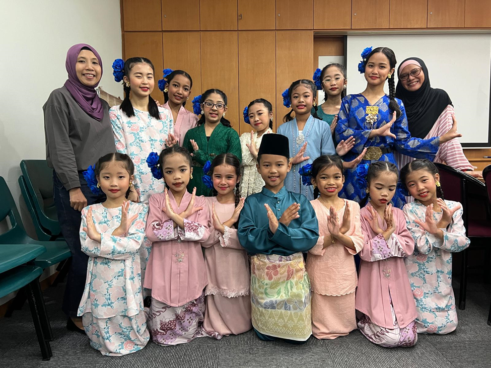 A group of children and adults in traditional Malay attire pose together, with many holding their hands in a dance gesture.