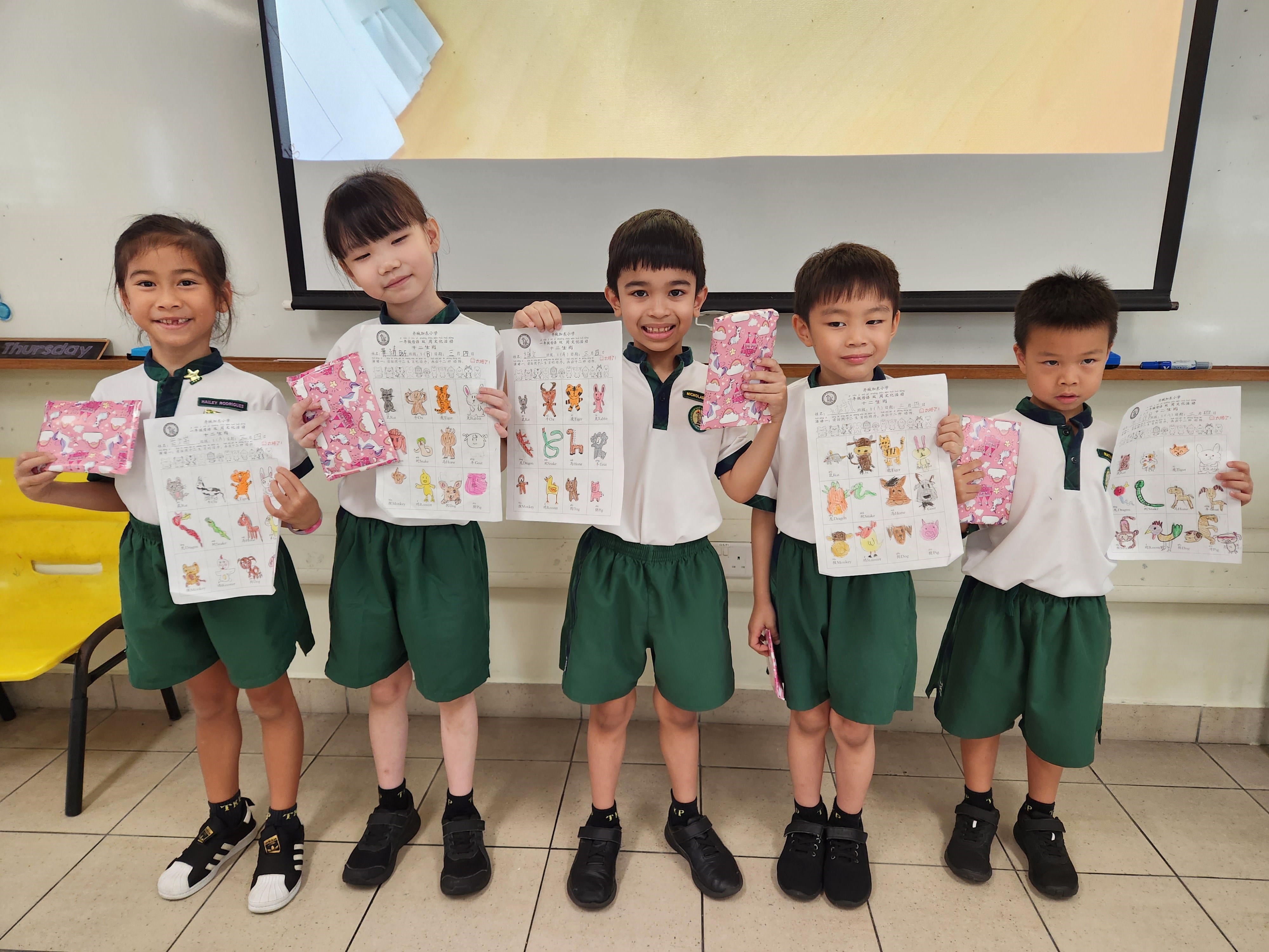Five young students in white and green uniforms hold up colorful animal drawing worksheets in a classroom.