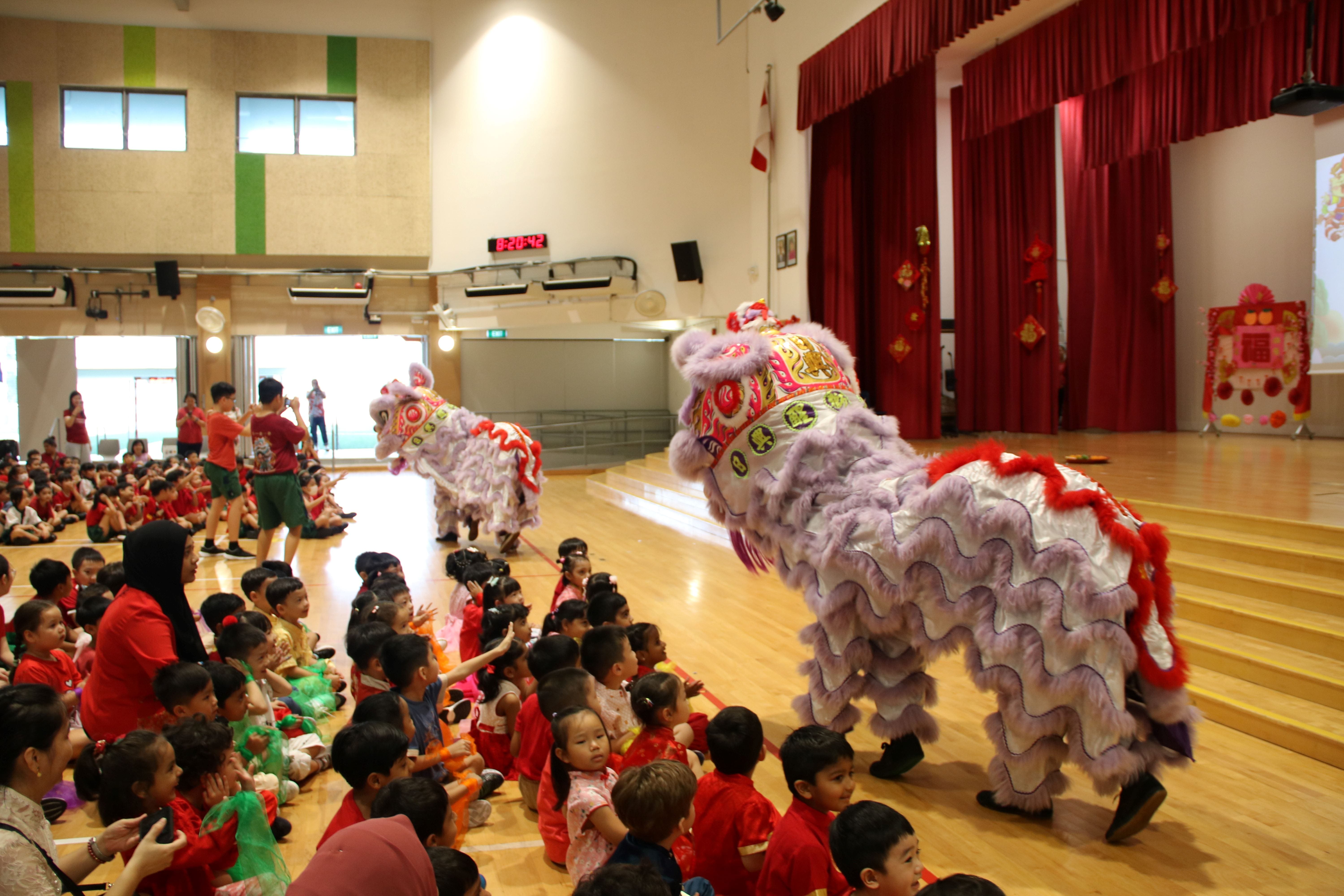 Two colorful lion dance costumes perform before a crowd of young children seated on a school hall floor.