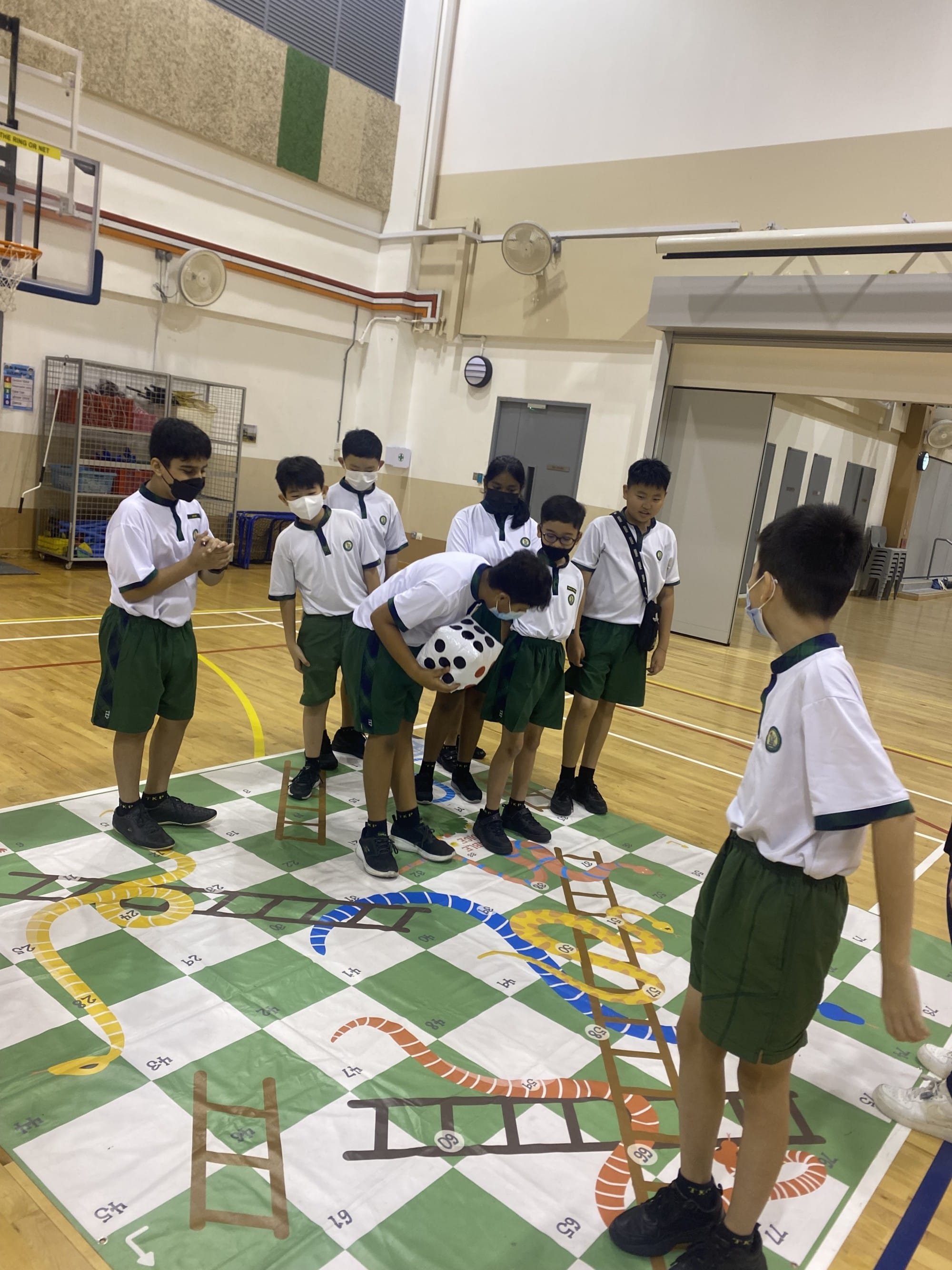 Students in school uniforms and masks play a life-size snakes and ladders game on a gym floor using a giant dice.