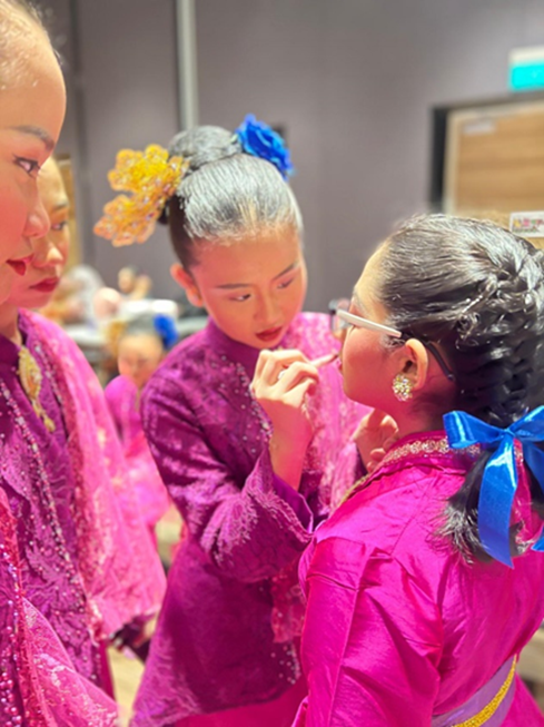 Three girls in bright pink traditional costumes with decorative hair accessories, one applying makeup to another.
