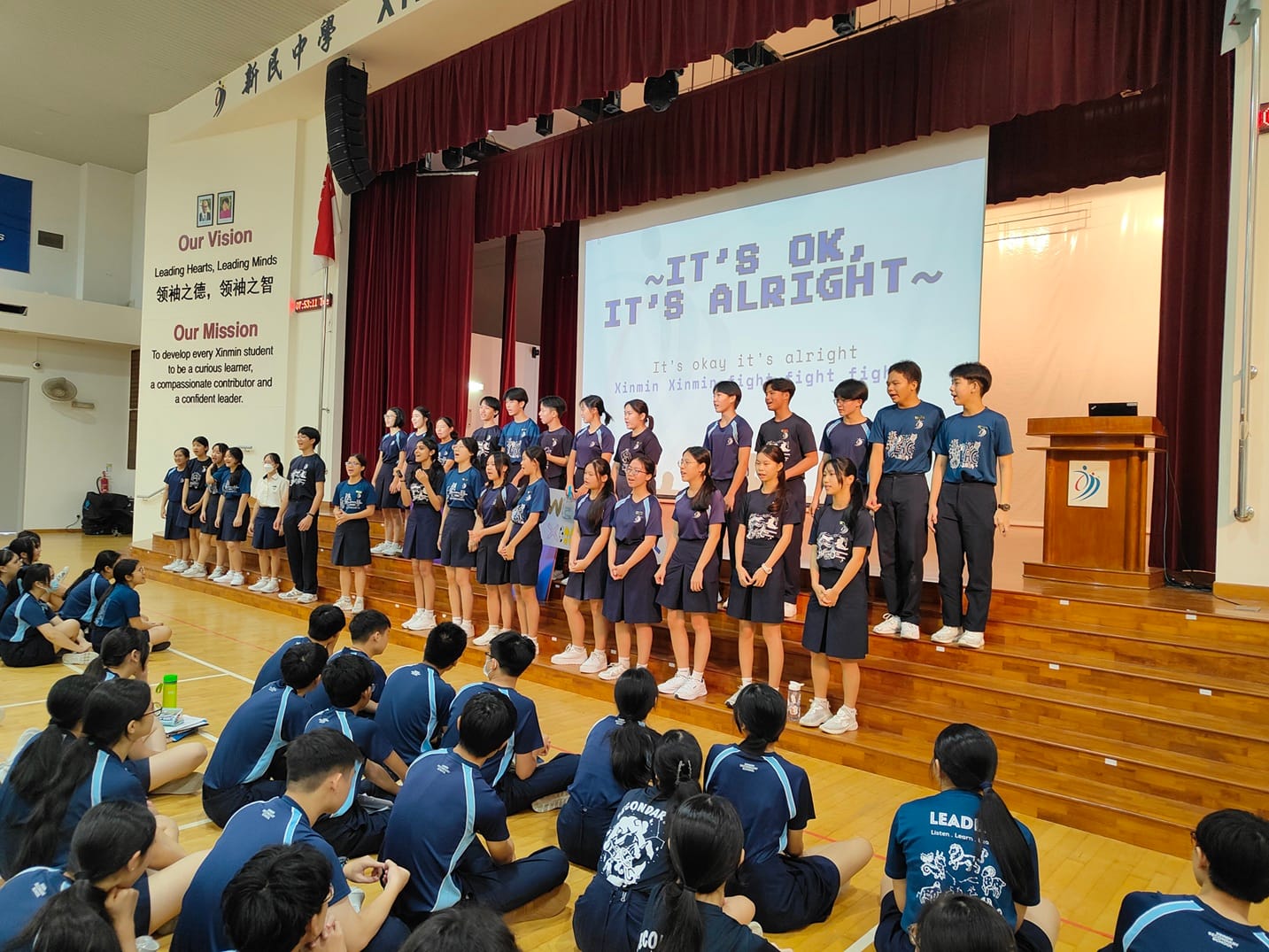 Students in navy uniforms perform on a school hall stage before a seated audience, with a screen showing "It's OK, It's Alright."