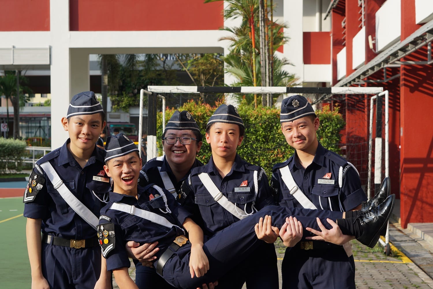 Five young students in navy uniforms with white sashes pose playfully outdoors, with three lifting one member horizontally.