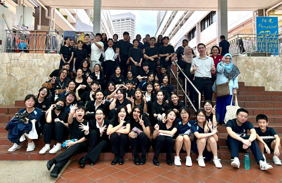 Large group of students mostly in black outfits posing on outdoor steps, making peace signs and excited expressions.