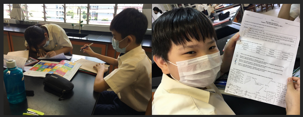 Two masked students study at a desk; a third masked boy holds up a completed worksheet.