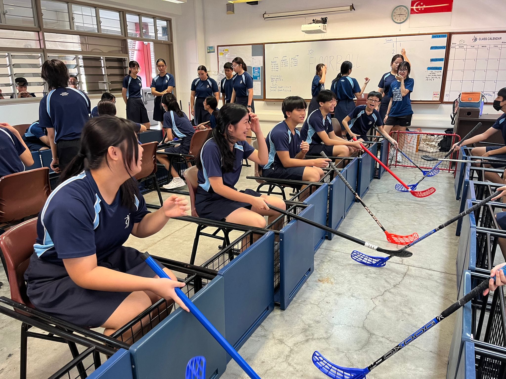 Students in navy uniforms sit at desks holding floorball sticks in a classroom with a whiteboard reading "Floorball."