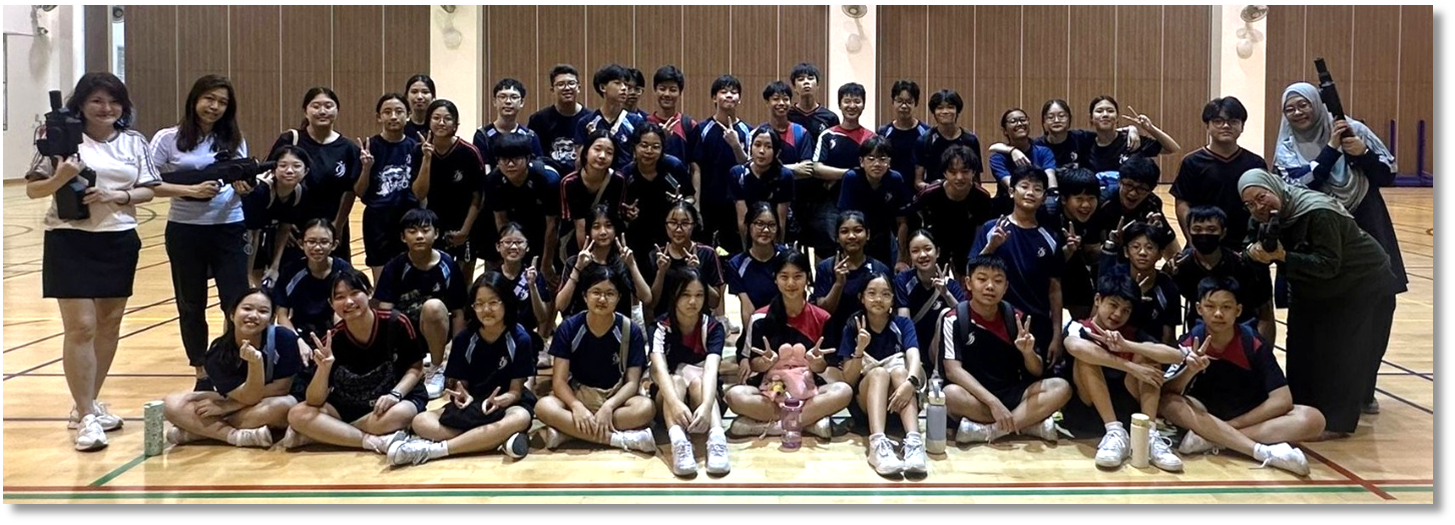 Large group of students in dark blue uniforms posing together in an indoor sports hall, many making peace signs.