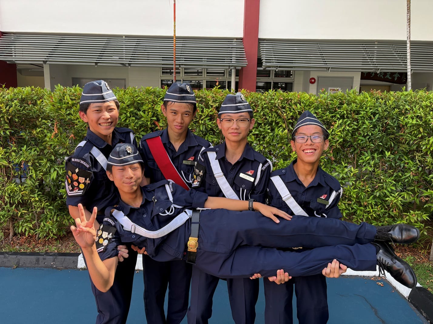 Five teenage boys in navy uniforms and caps pose outdoors, with three lifting one boy horizontally.