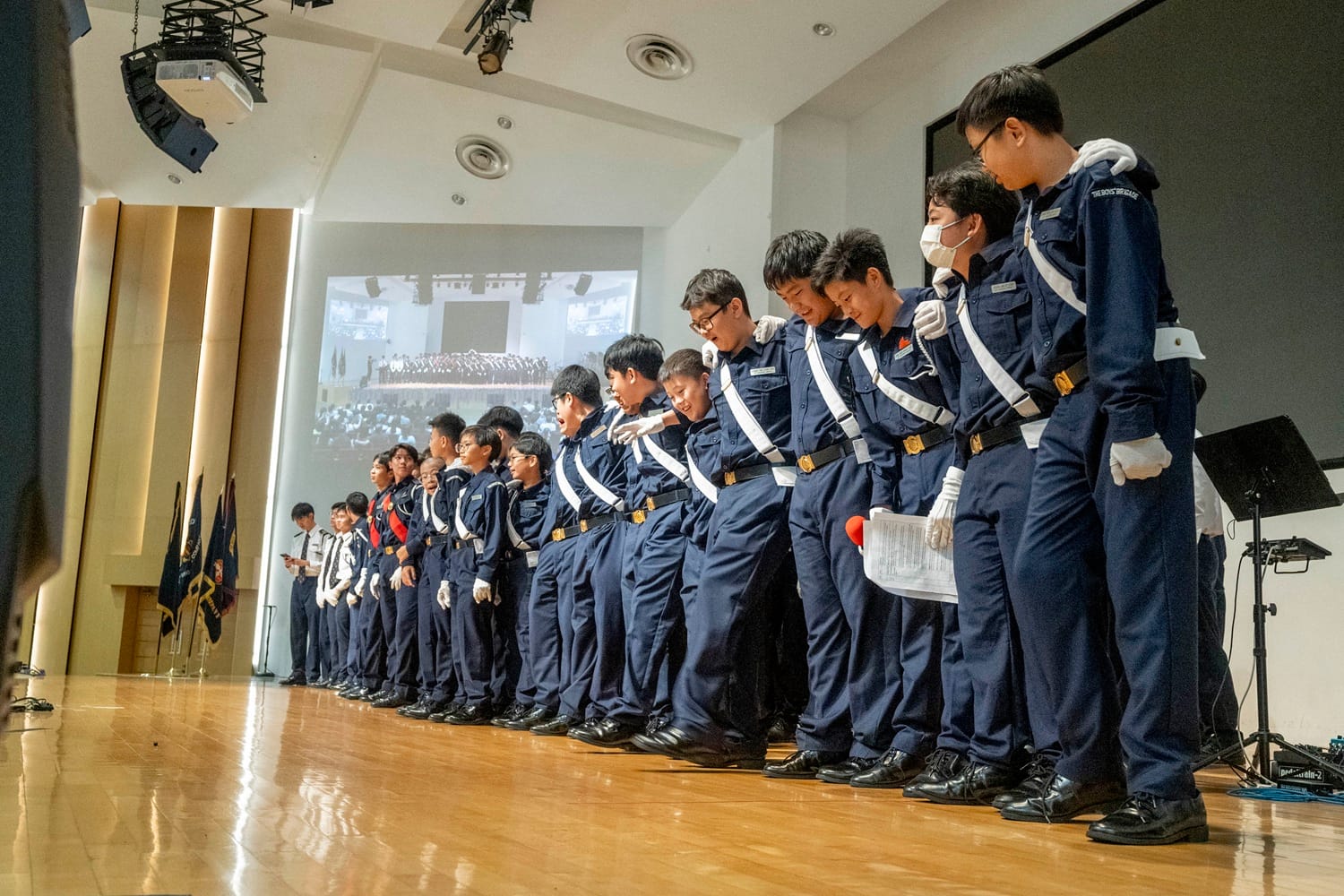 A row of young boys in navy blue uniforms with white belts and gloves stand on a wooden stage during a ceremony.