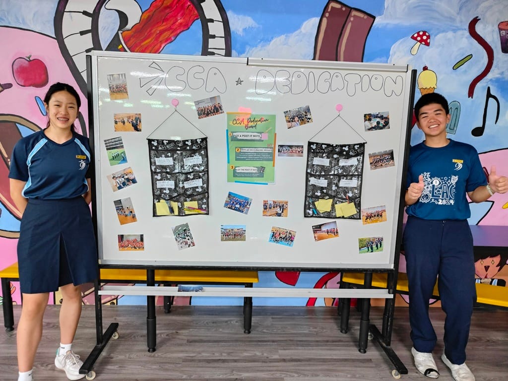 Two students in navy uniforms stand beside a whiteboard labeled CCA Dedication, covered with photos and notes.