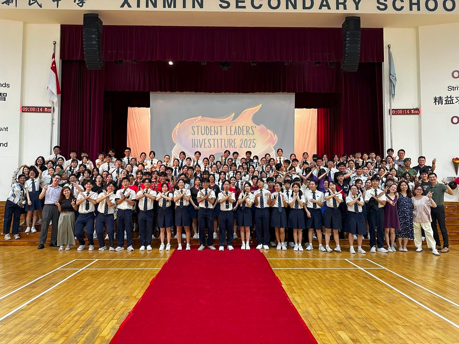 Large group of students and staff posing at Xinmin Secondary School Student Leaders Investiture 2025 on a stage.