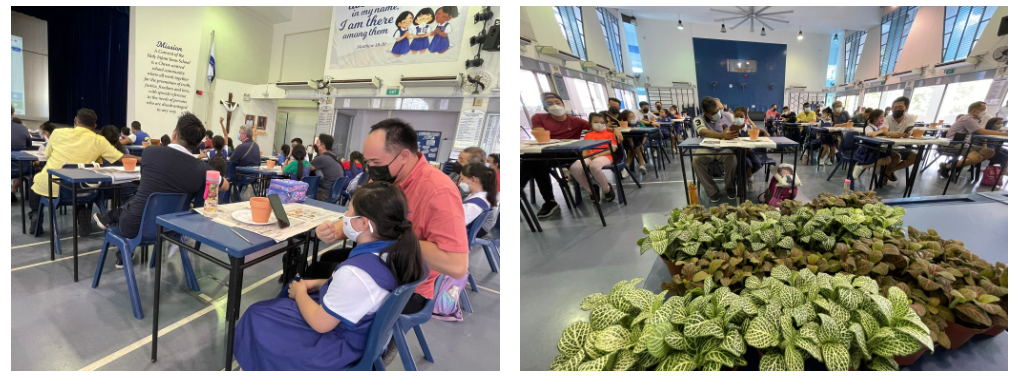 Adults and children wearing masks sit at tables in a school hall; potted plants are visible in the foreground.