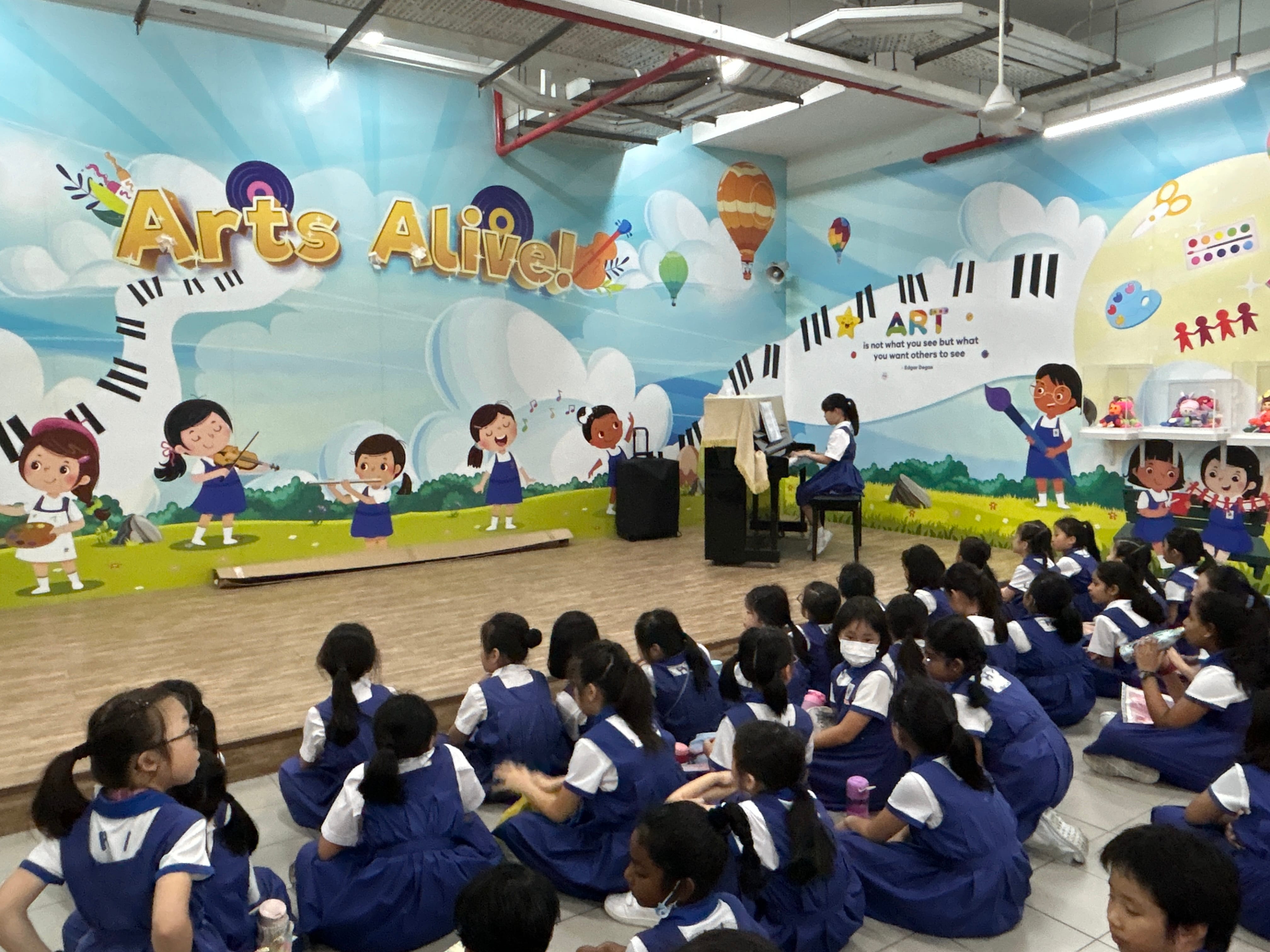 Young students in blue uniforms sit on the floor watching a girl play piano on a stage with an Arts Alive mural behind her.