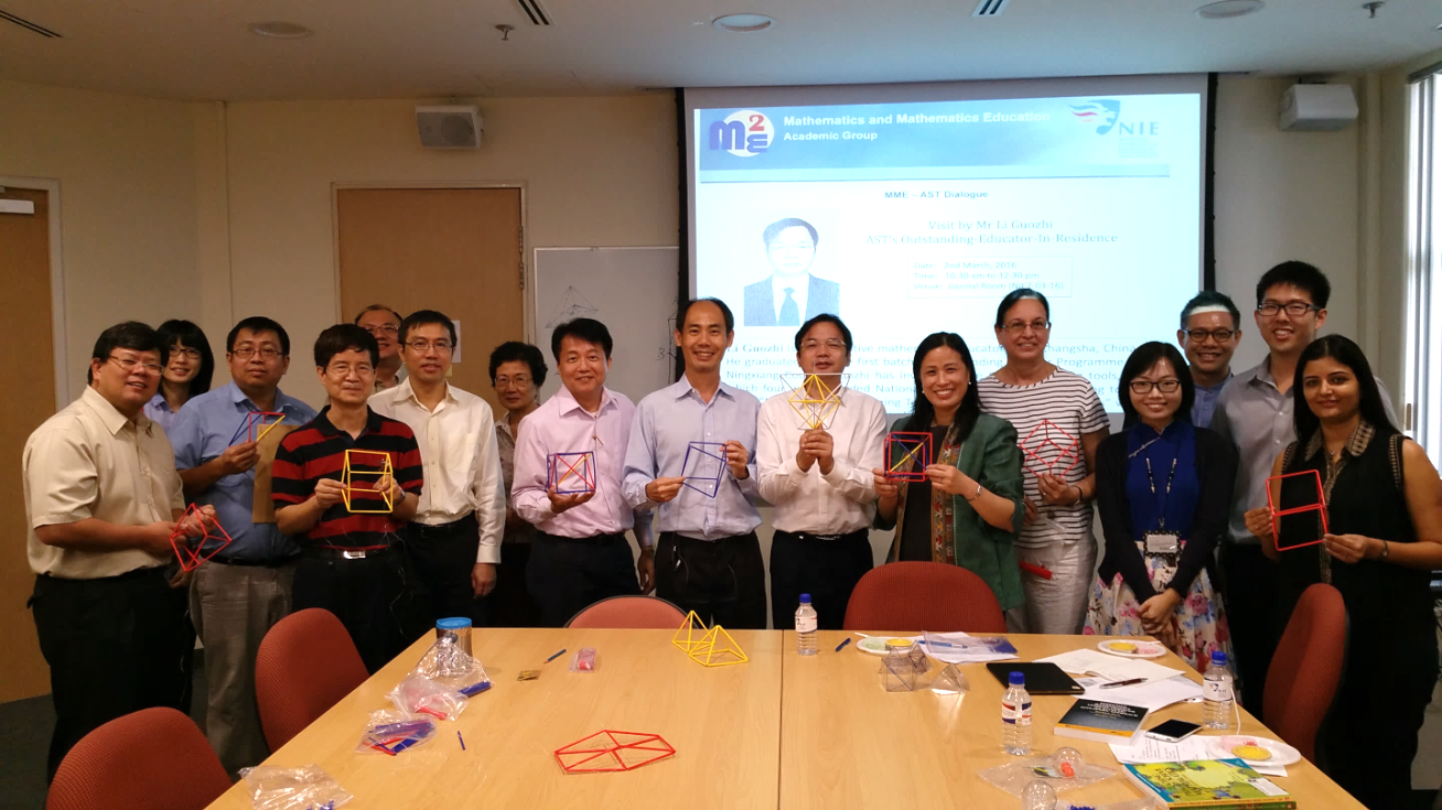 A group of people stand in a conference room holding colorful geometric wire models, with a mathematics education presentation on screen behind them.