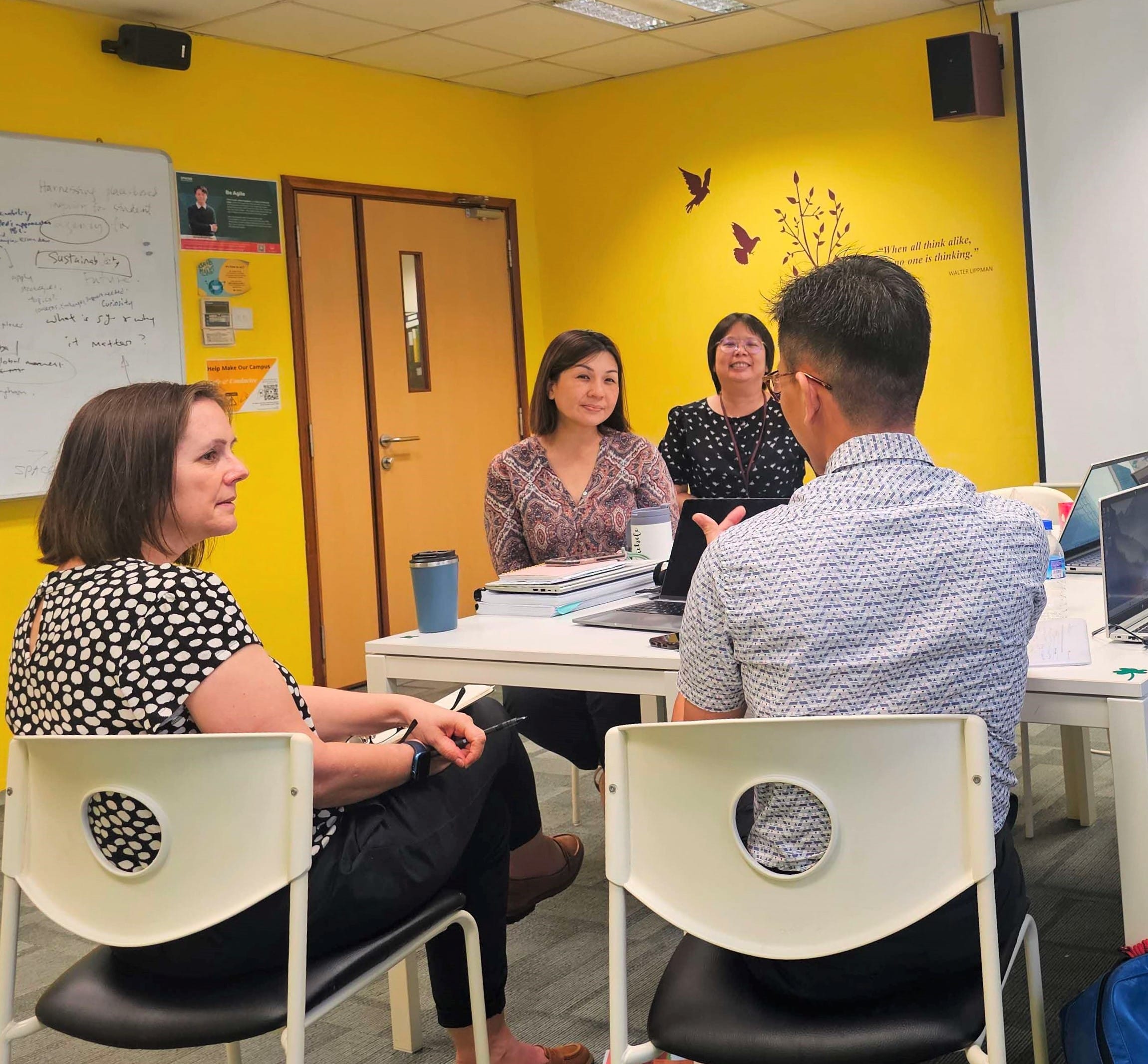 Four people sit around a white table in a yellow-walled room, engaged in conversation near a whiteboard.