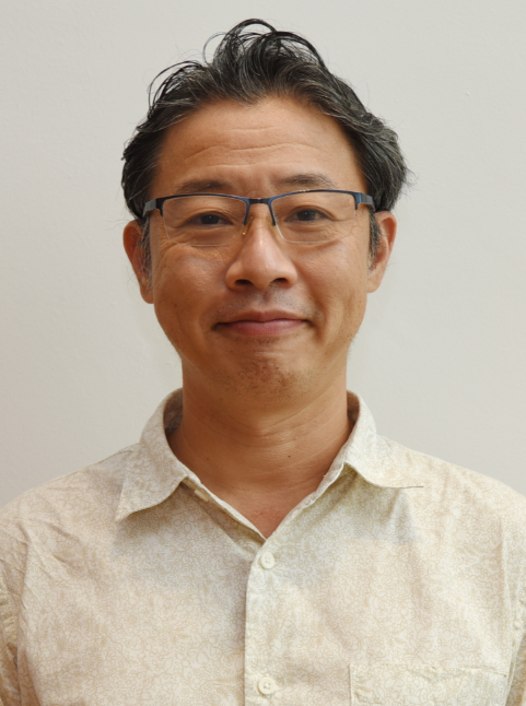 Middle-aged man with dark hair and glasses wearing a light beige button-up shirt, smiling slightly against a plain wall.