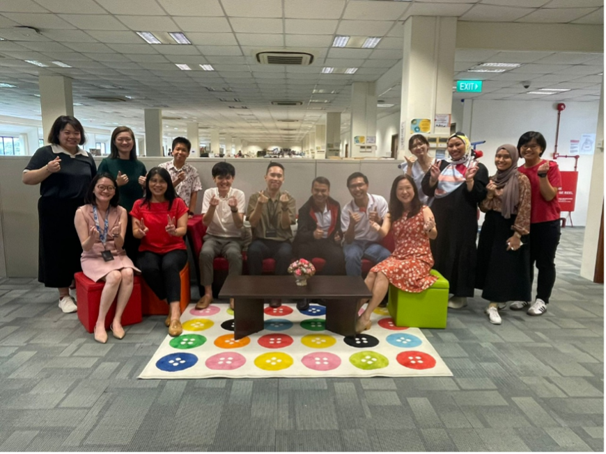 A group of about 14 people posing and smiling in an office lounge area with a colorful button-print rug.