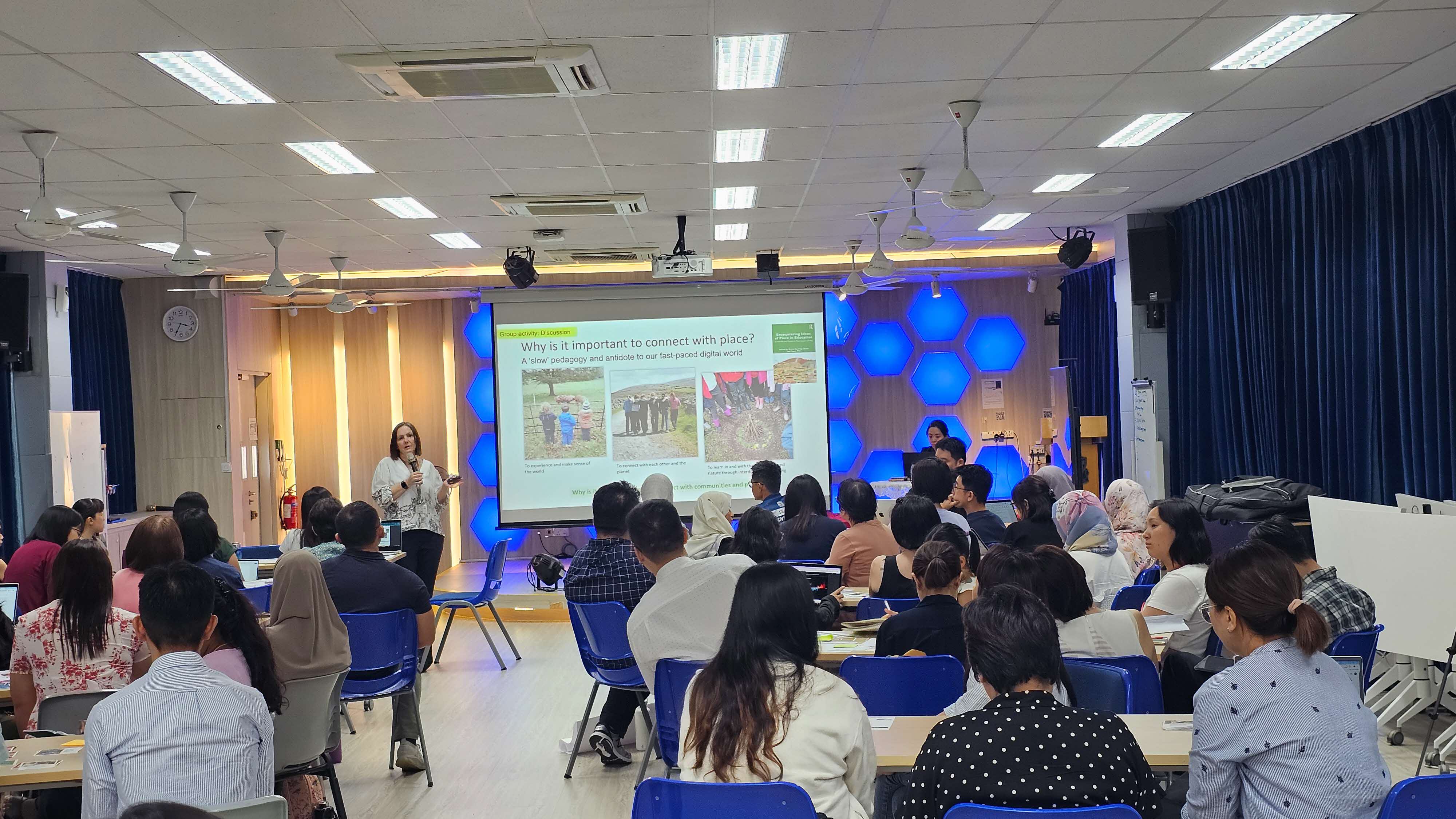 A woman presents to a seated audience in a conference room, with a projected slide reading "Why is it important to connect with place?"