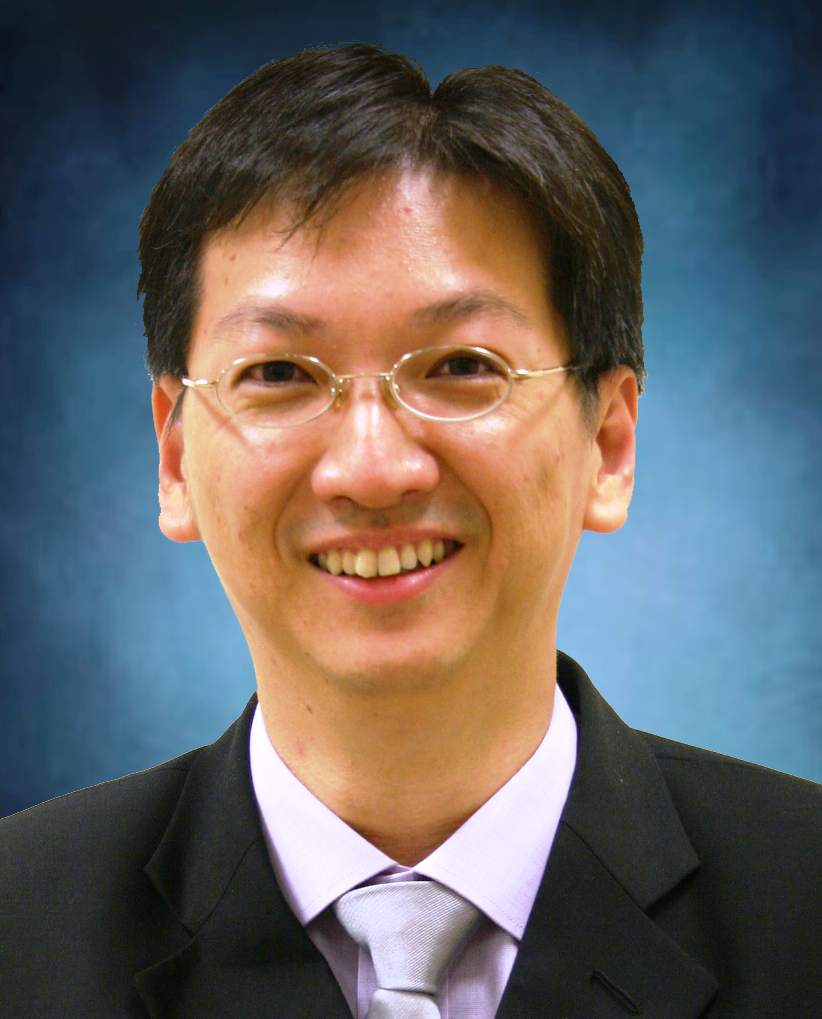 A smiling man with glasses, dark hair, wearing a black suit, light purple shirt, and gray tie against a blue background.