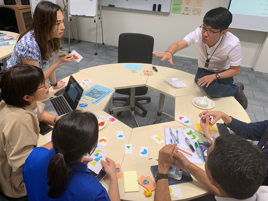 Five people sit around a curved table covered with colorful cards and sticky notes, discussing in a classroom setting.