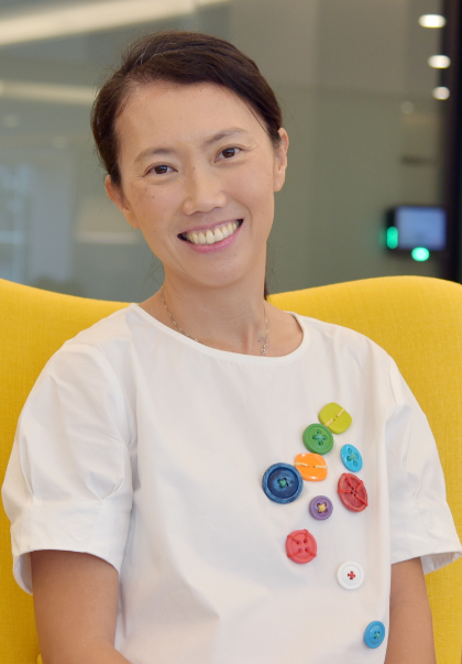 A smiling woman in a white top with colorful buttons pinned on it, seated in a yellow chair.