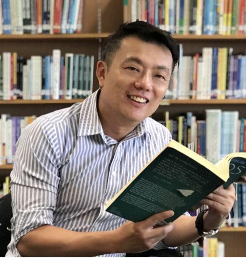 A smiling man in a striped shirt holds an open book in front of shelves filled with colorful books.