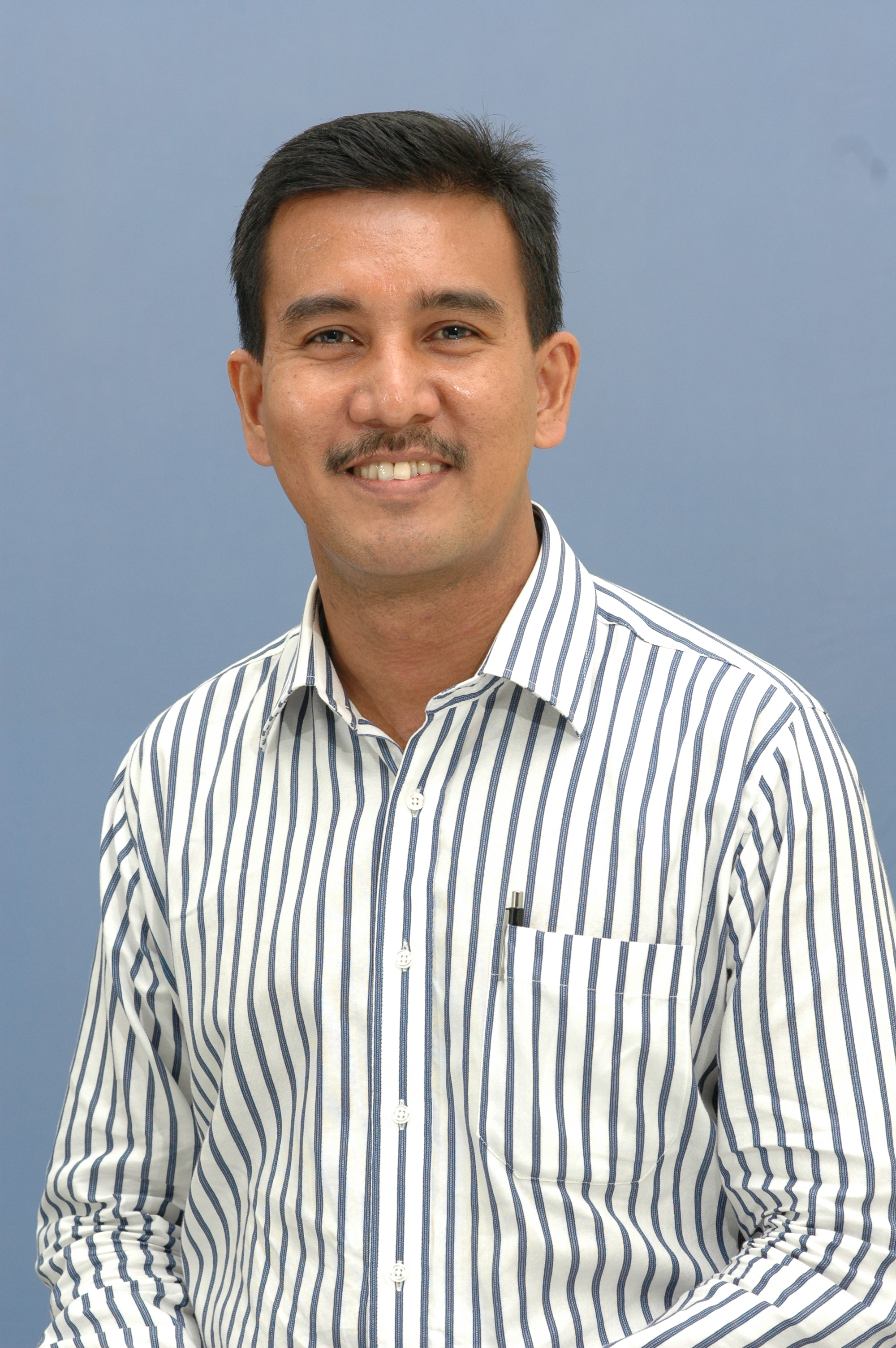 A smiling man with a mustache wearing a white and blue striped shirt against a light blue background.