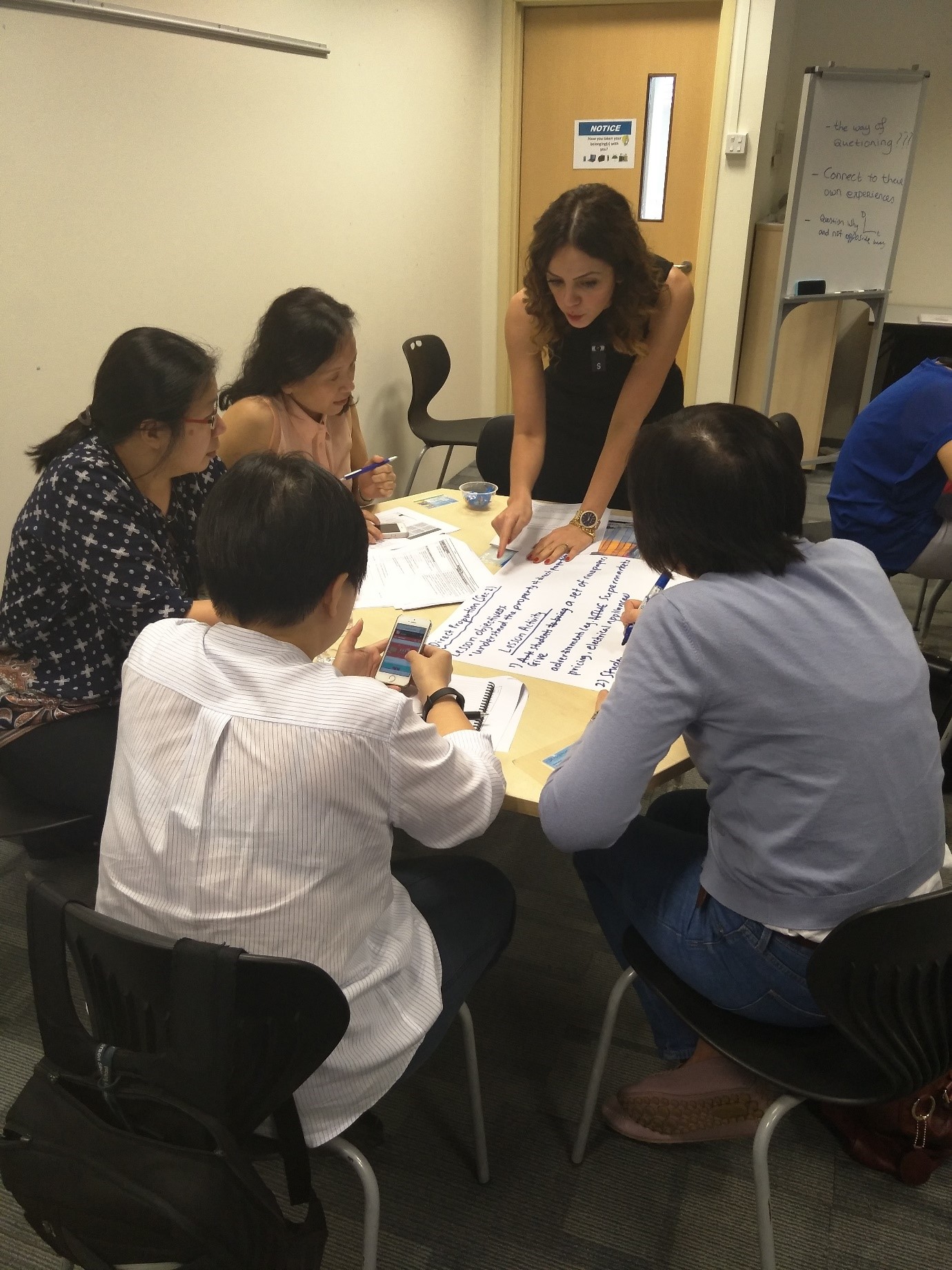Five people gather around a table covered in papers, with one woman standing and pointing at a large handwritten sheet.