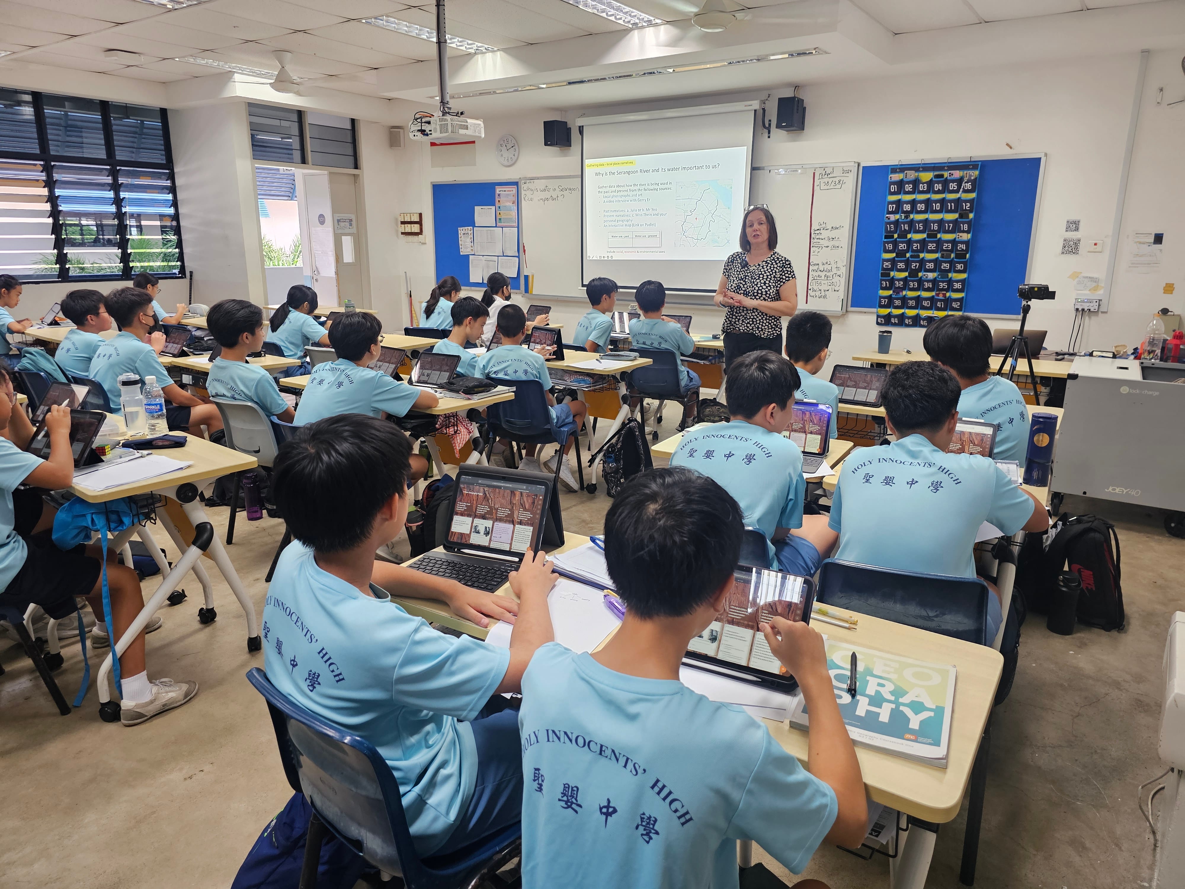 A teacher stands at a projector screen in a classroom full of students in light blue Holy Innocents' High uniforms using laptops and tablets.