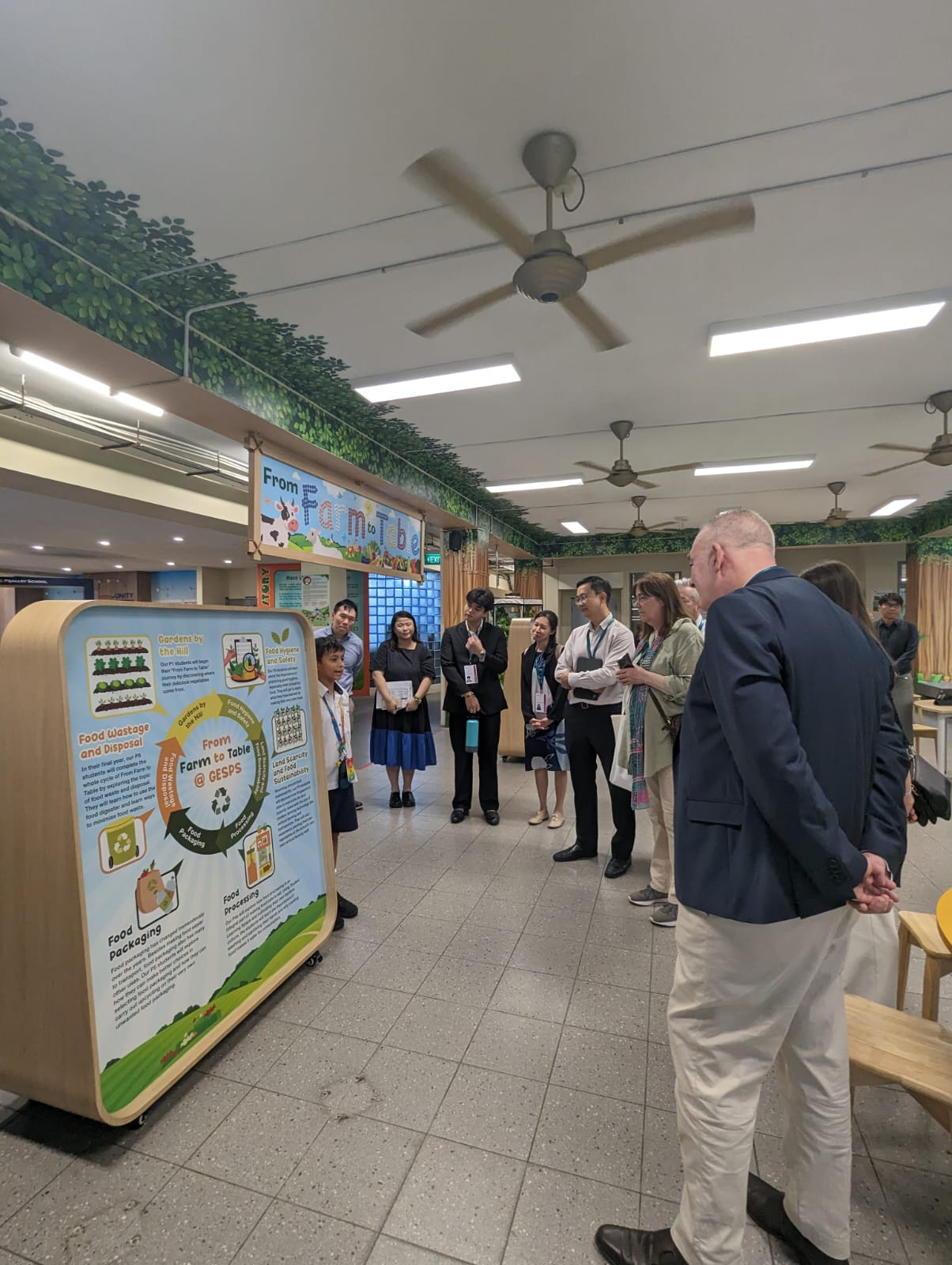 A group of people stands indoors viewing a colorful Farm to Table educational display board.