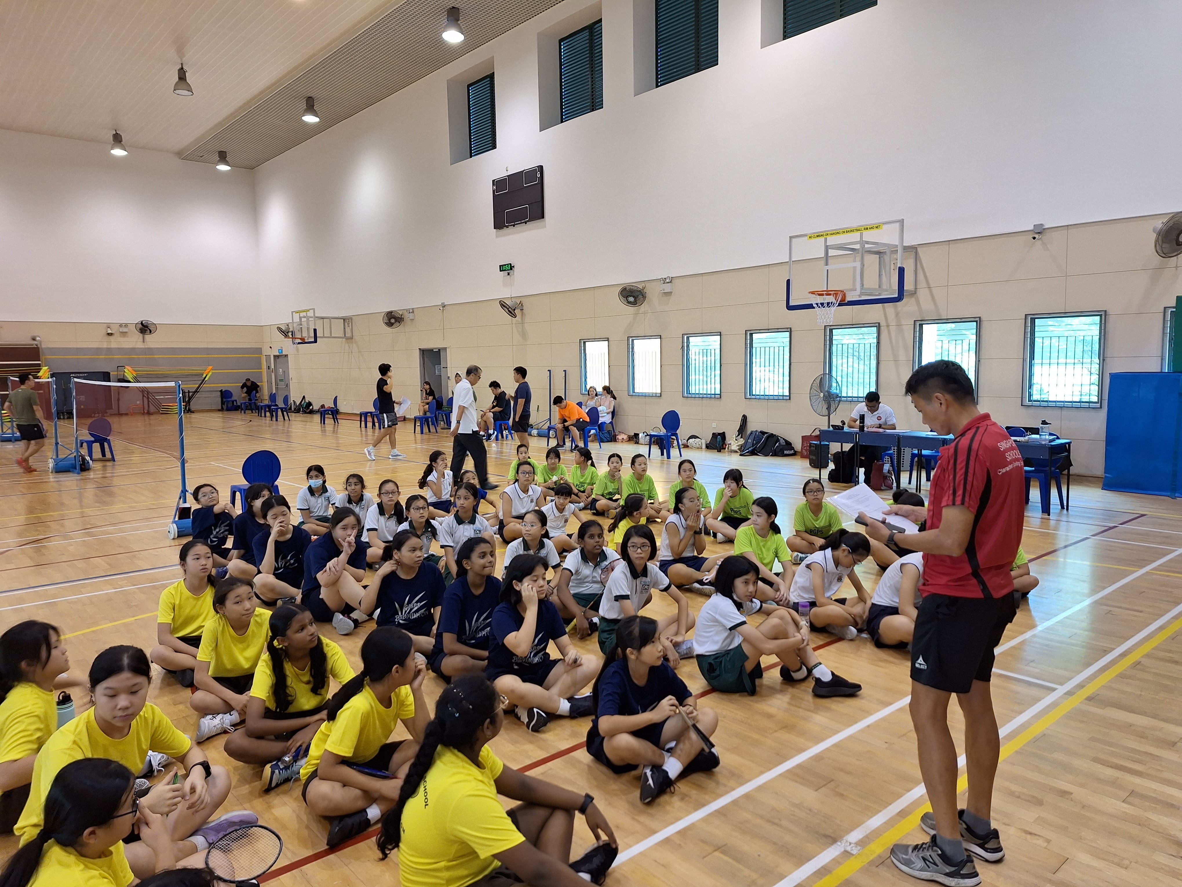 A coach in red addresses a large group of students seated on an indoor sports hall floor during a badminton event.