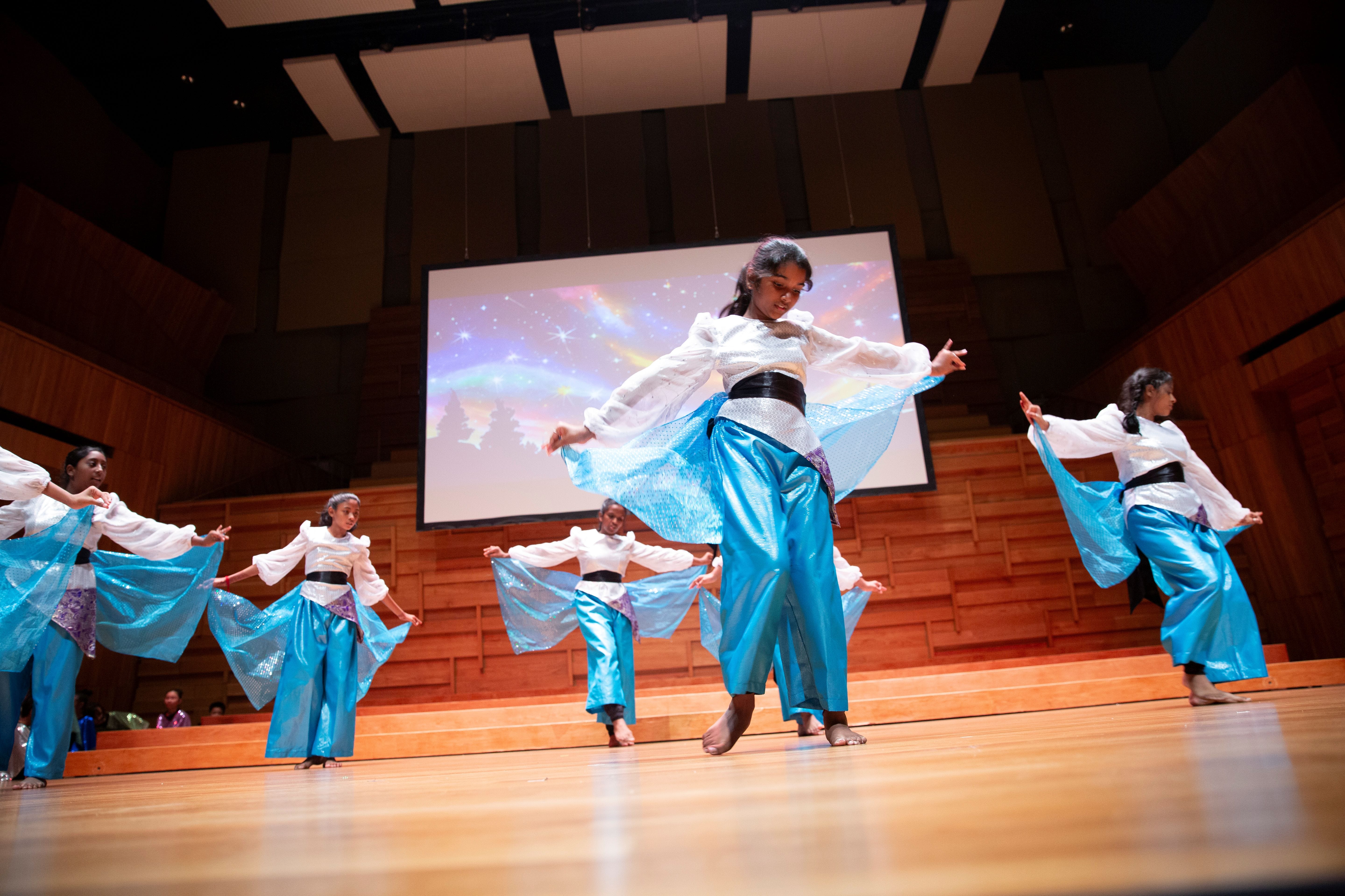 Several girls in white tops and blue satin pants perform a dance on a wooden stage with a colorful screen behind them.