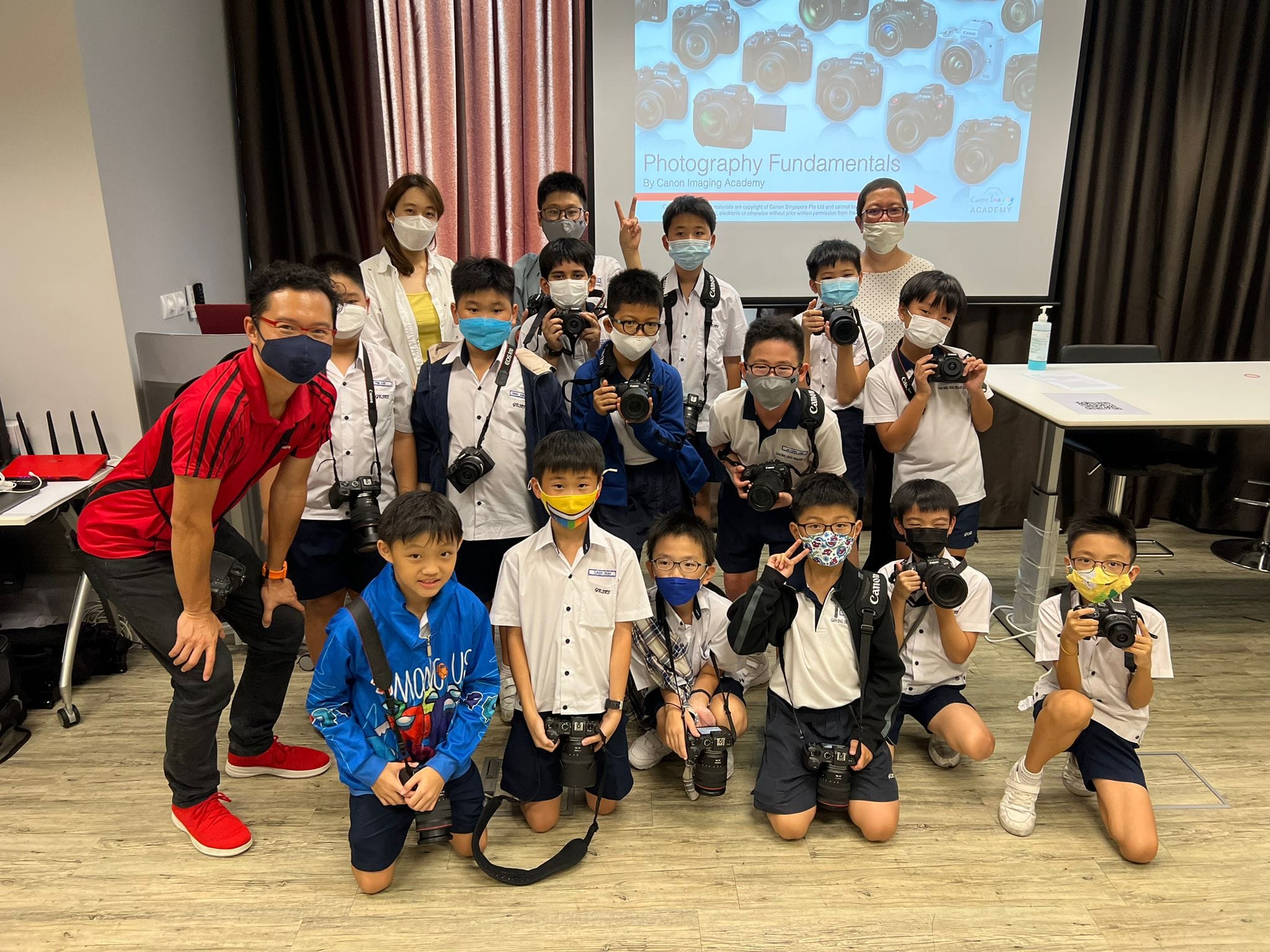 A group of masked children and adults pose with cameras at a Photography Fundamentals class by Canon Imaging Academy.