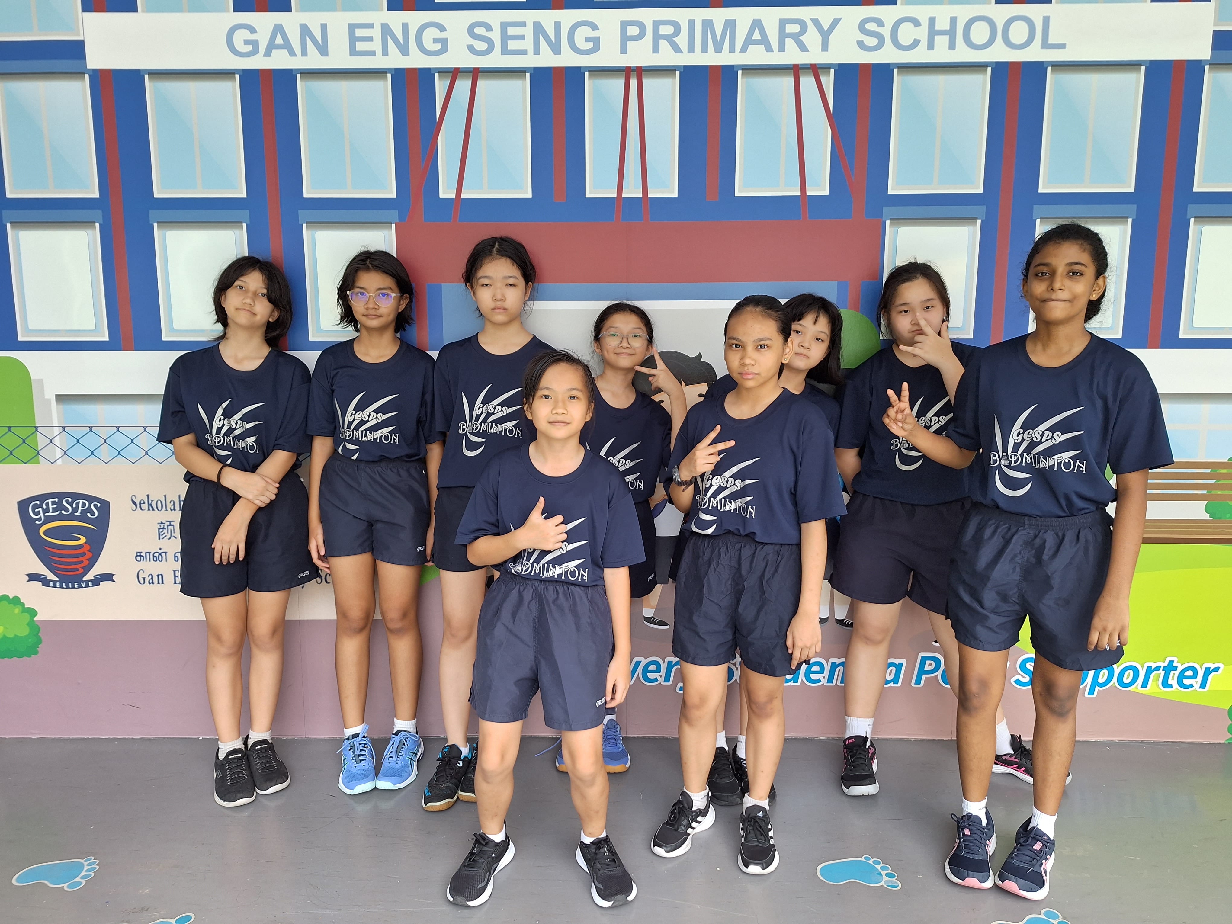 Nine students in navy GESPS Badminton uniforms pose in front of a Gan Eng Seng Primary School mural.