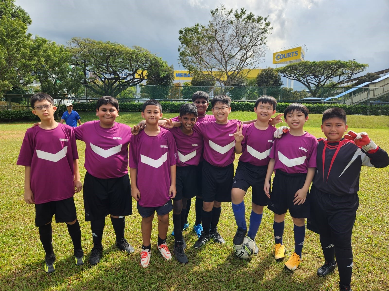 Nine boys in purple football jerseys pose together on a grass field, with trees and a Courts sign behind them.