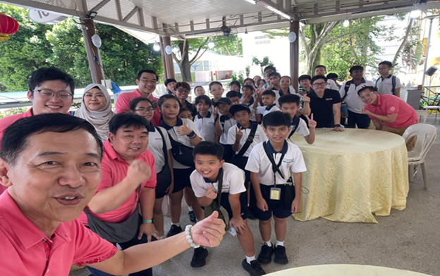 Group of adults and children in school uniforms posing with thumbs up under an outdoor pavilion.