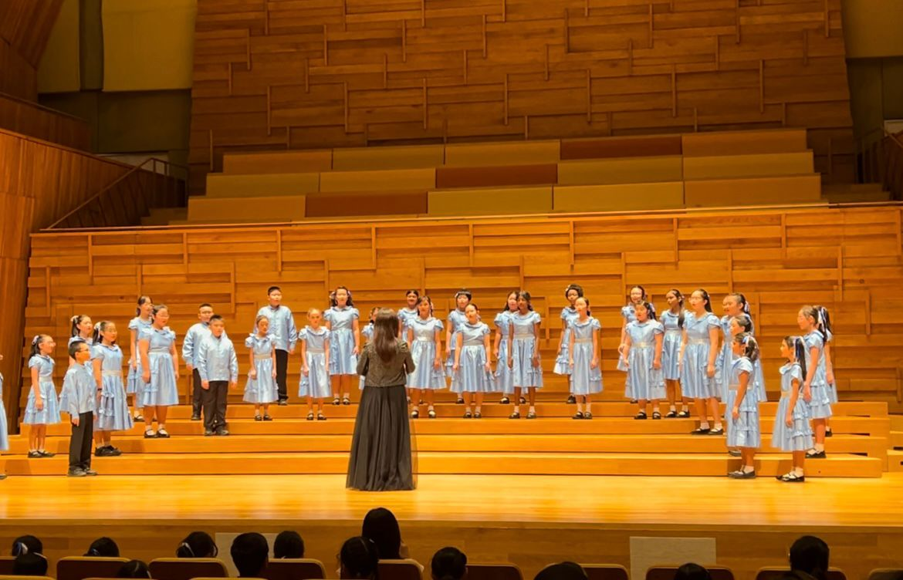 A conductor leads a children's choir in light blue uniforms performing on a wooden concert hall stage.