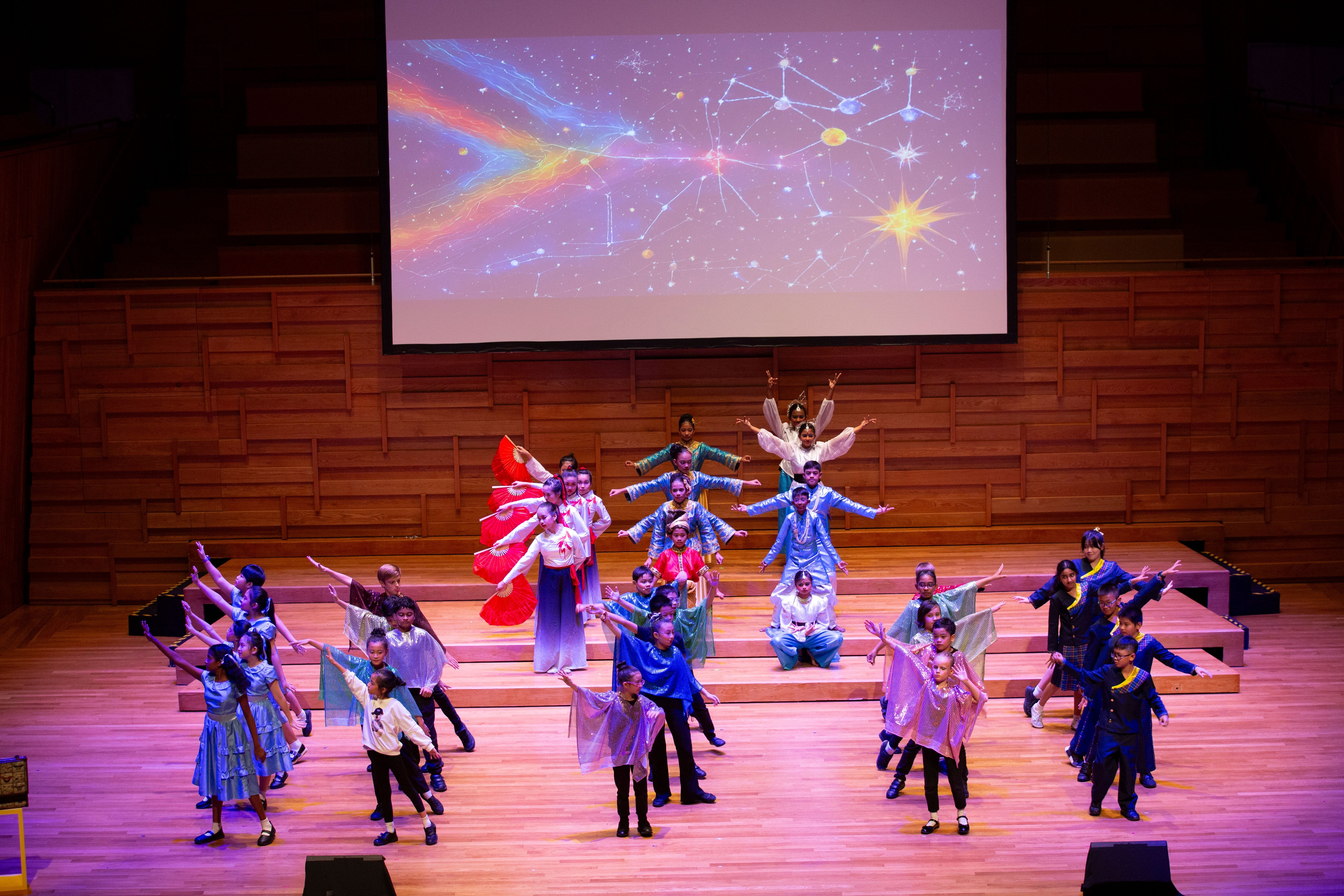 Children in colorful cultural costumes perform a dance on a wooden stage with a starry constellation backdrop.