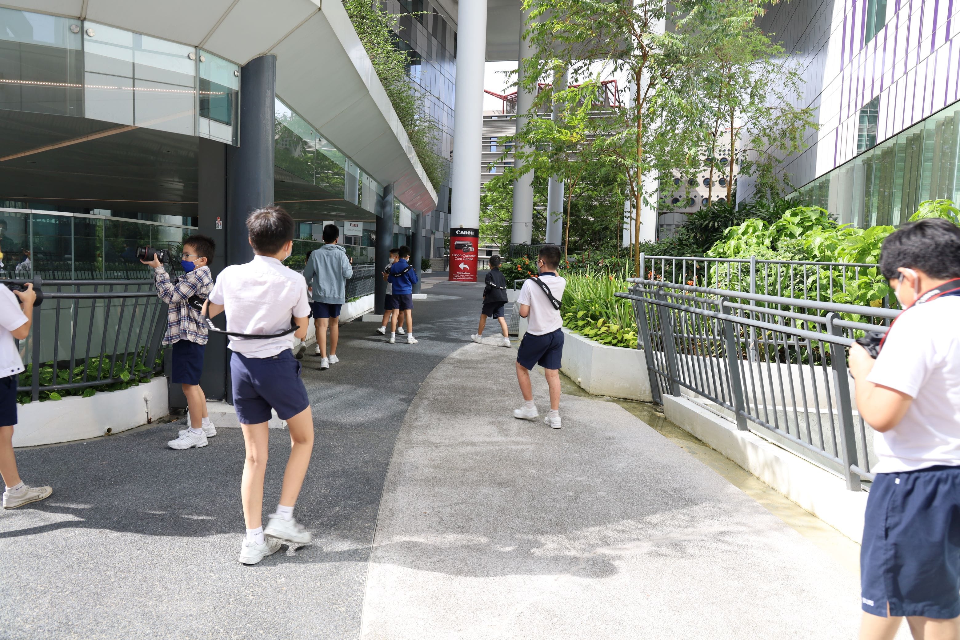 Students in school uniforms walk and take photos with cameras outside a modern glass building with green plants.