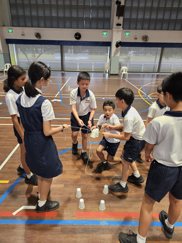 Six uniformed school children stand in a circle on a gym floor, cooperating to lift a cup using strings.
