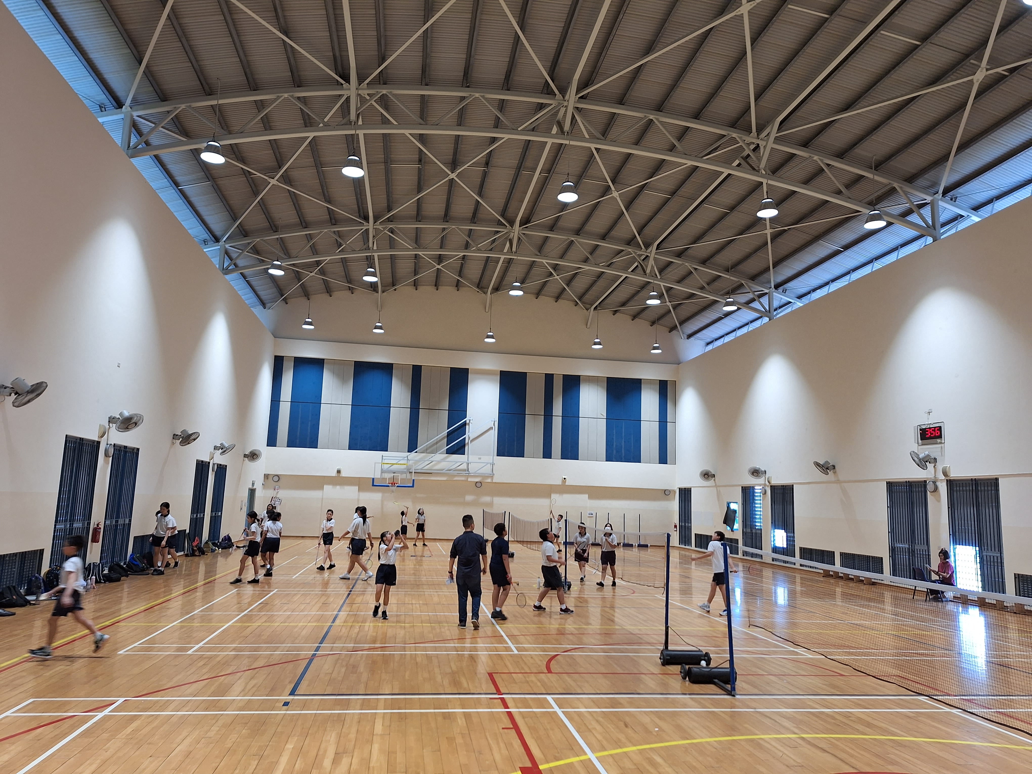 Students in white and black uniforms play badminton on a wooden indoor court with high arched metal ceiling.