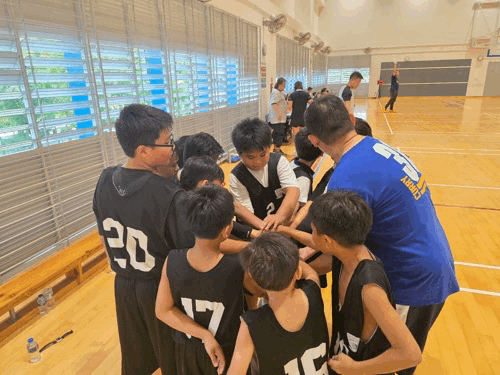 Young basketball players in black jerseys huddle together on an indoor court, stacking hands in a team cheer.
