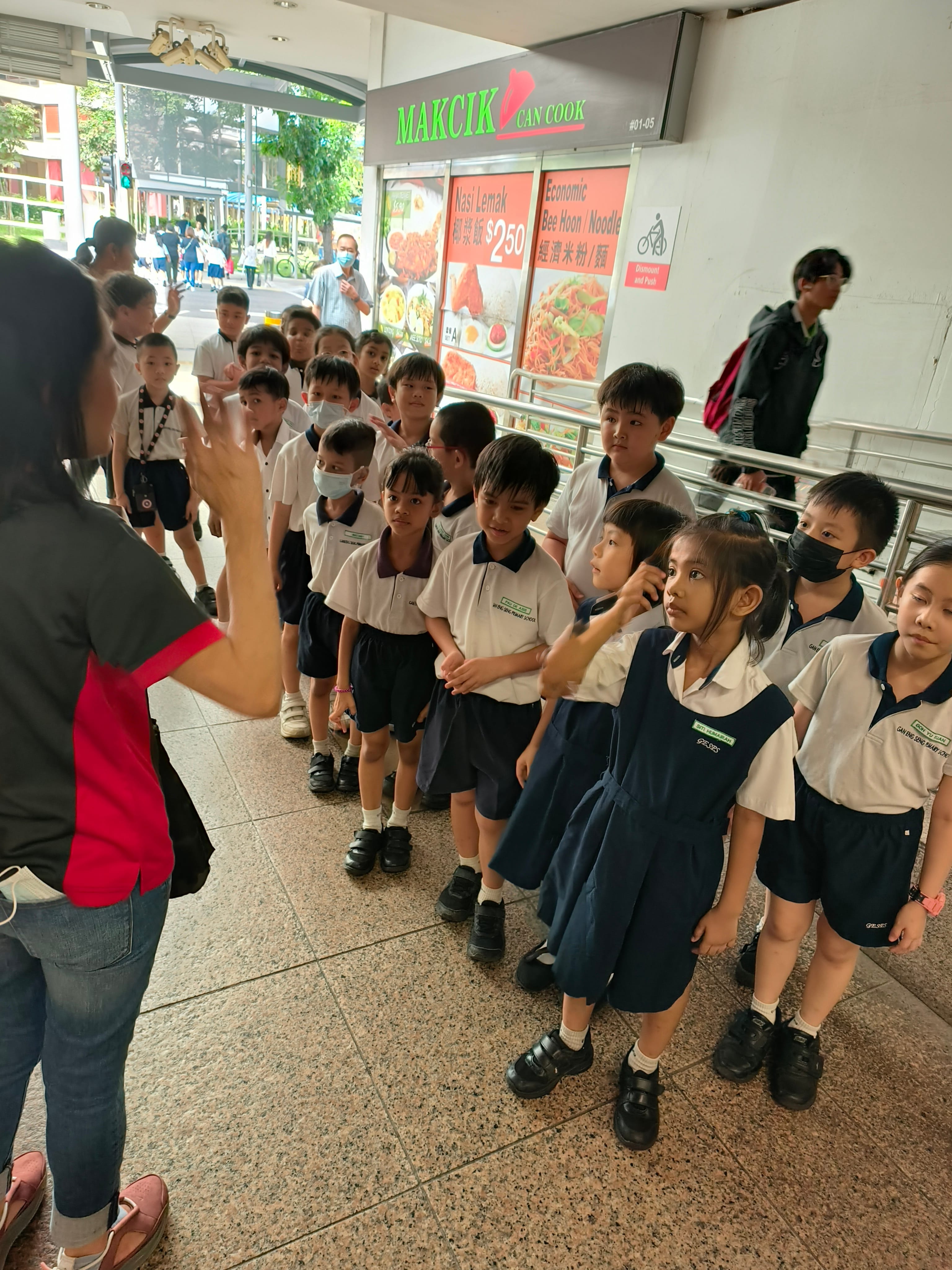 A teacher gestures to a group of young students in school uniforms gathered outside a food stall called Makcik Can Cook.